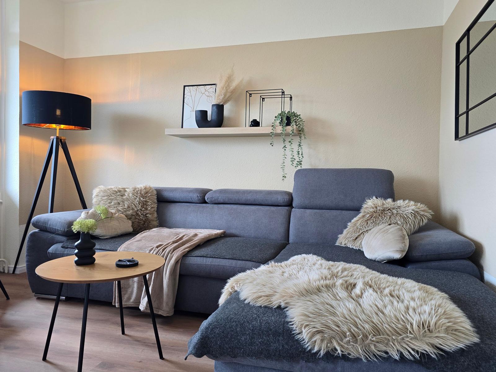 Ein grauer Sofa mit Felldecke und Kissen. Holztisch und Stehlampe daneben.
