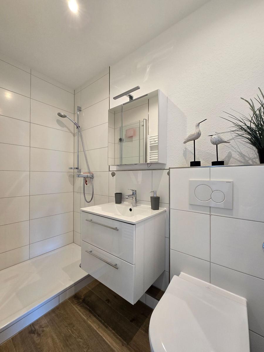 Badezimmer mit Dusche, Waschbecken und Toilette. Weiße Fliesen und Holzboden.