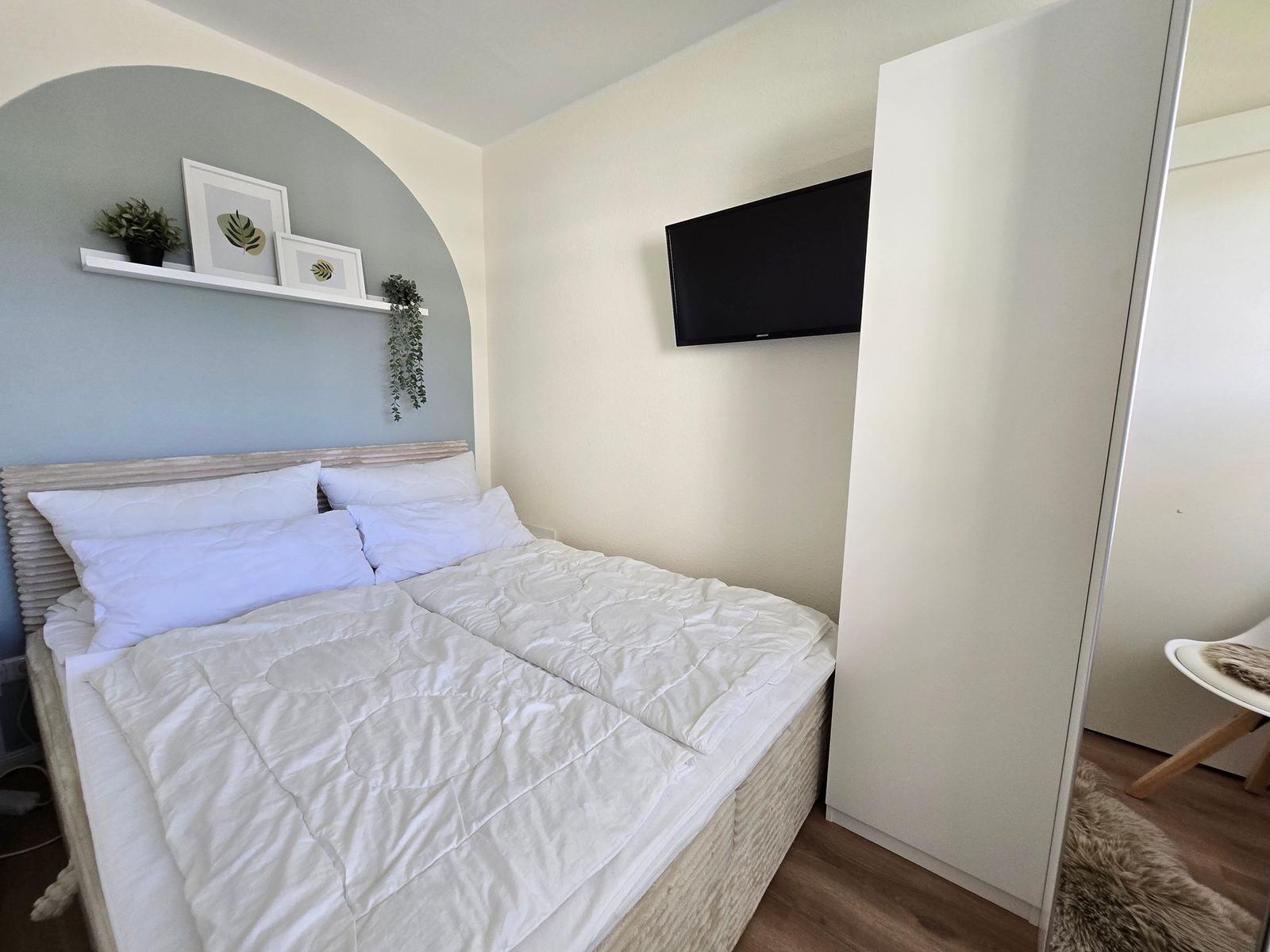 Schlafzimmer mit Bett, Wand-TV und Schrank. Wand mit Regal und Pflanzen.