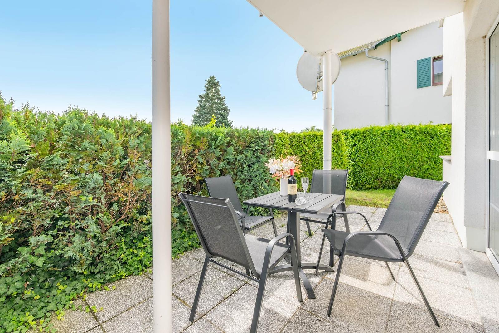 Terrasse mit Tisch, vier Stühlen, Blumen und Weinflasche. Umgeben von grünen Hecken.
