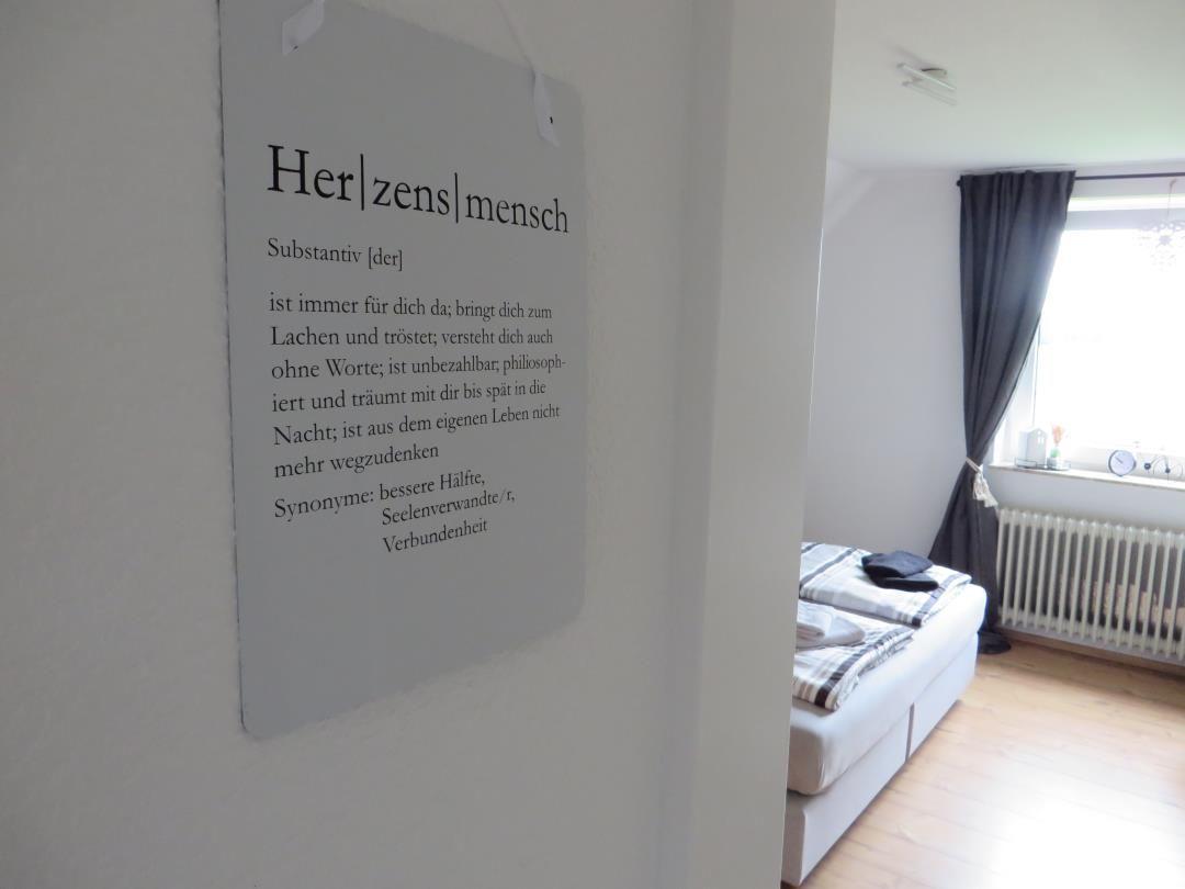 Zimmer mit Bett, Fenster und Wandtafel mit Text.