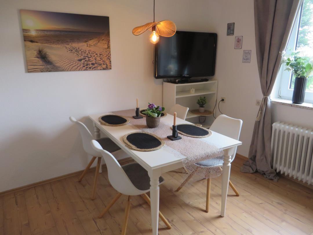 Esszimmer mit weißem Tisch, vier Stühlen und Fernseher an der Wand.