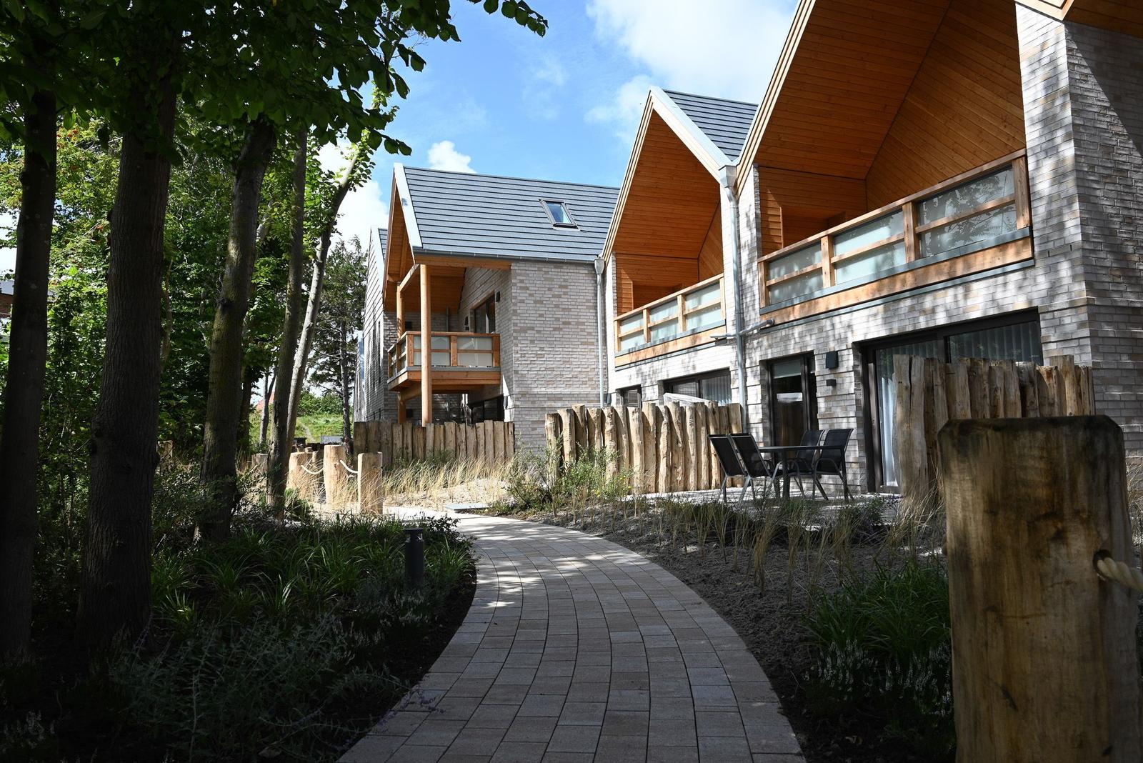 Wohnhaus mit Holzveranda und Steinmauer im Wald. Pflasterweg führt durch Grünanlagen.