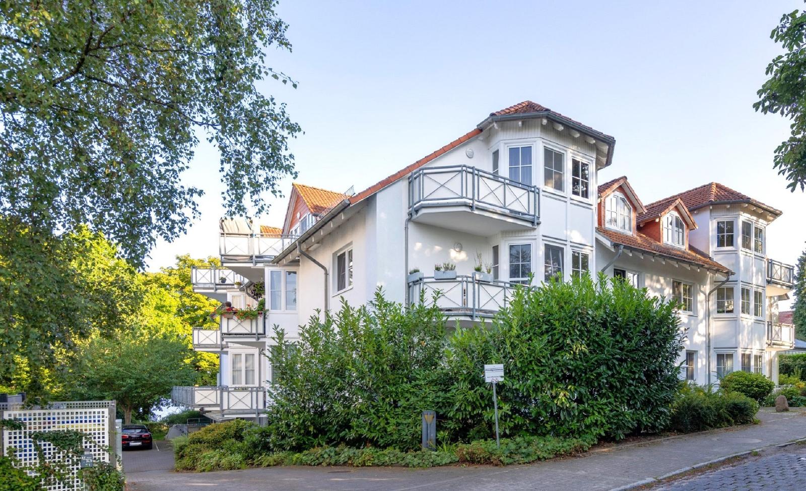 Weißes Mehrfamilienhaus mit Balkonen und rotem Dach unter Bäumen