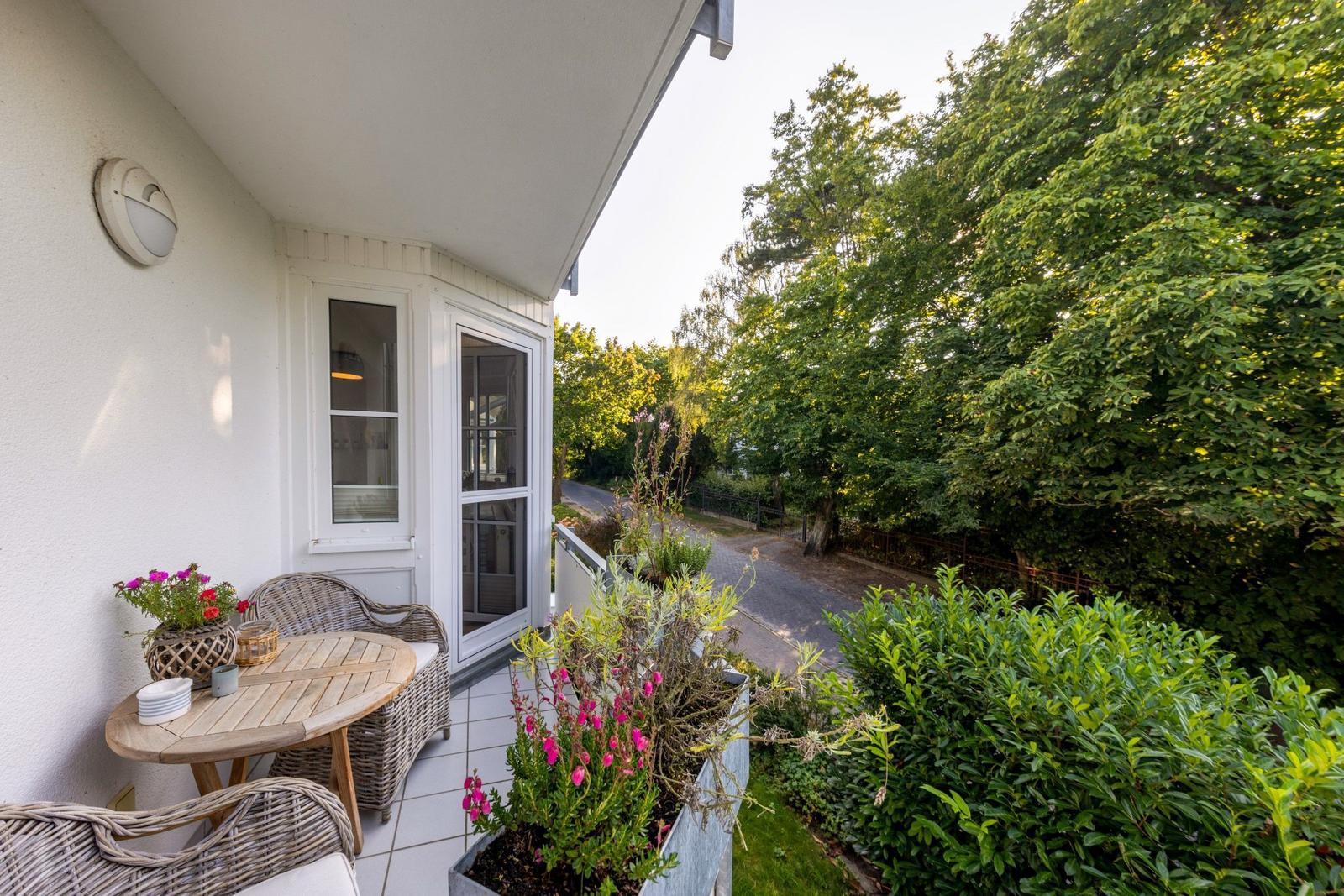 Terrasse mit Tisch, Stühlen und Pflanzen. Blick auf grüne Bäume und einen Weg.