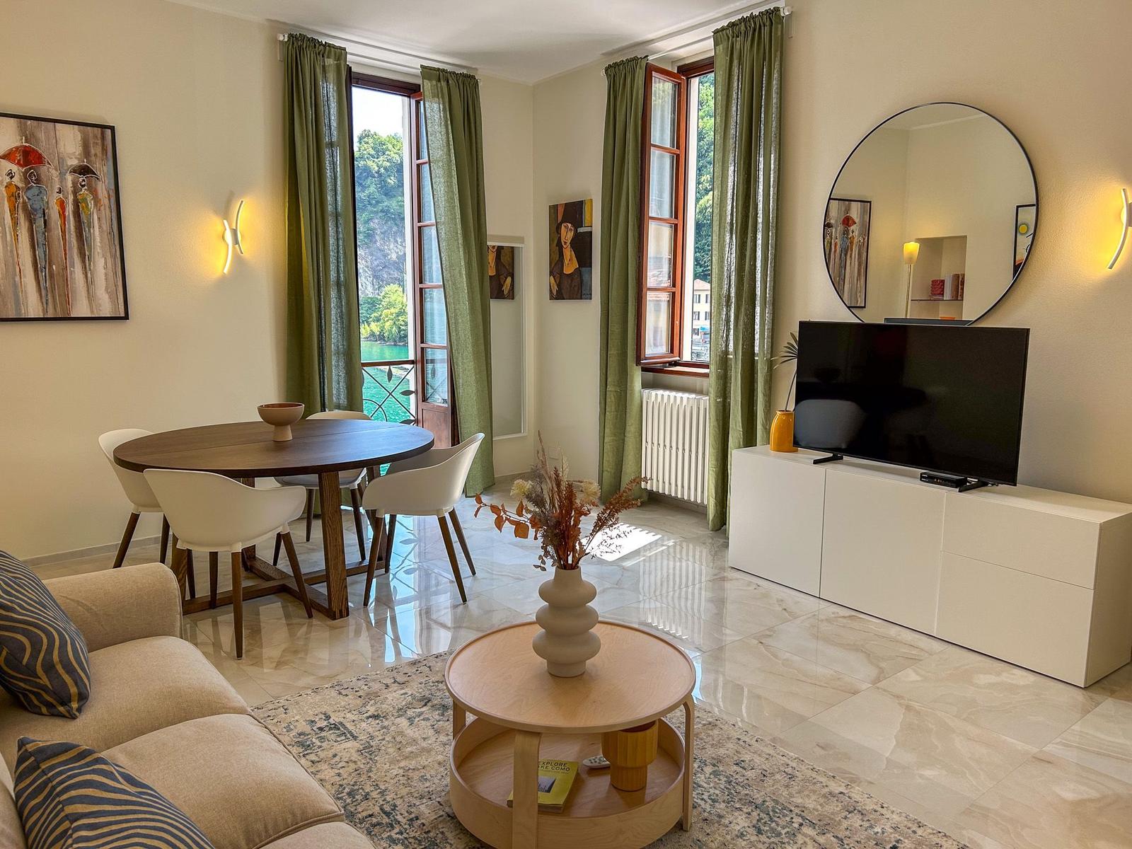 Living and dining area with TV, table, and sofa. Green curtains and windows with water view.