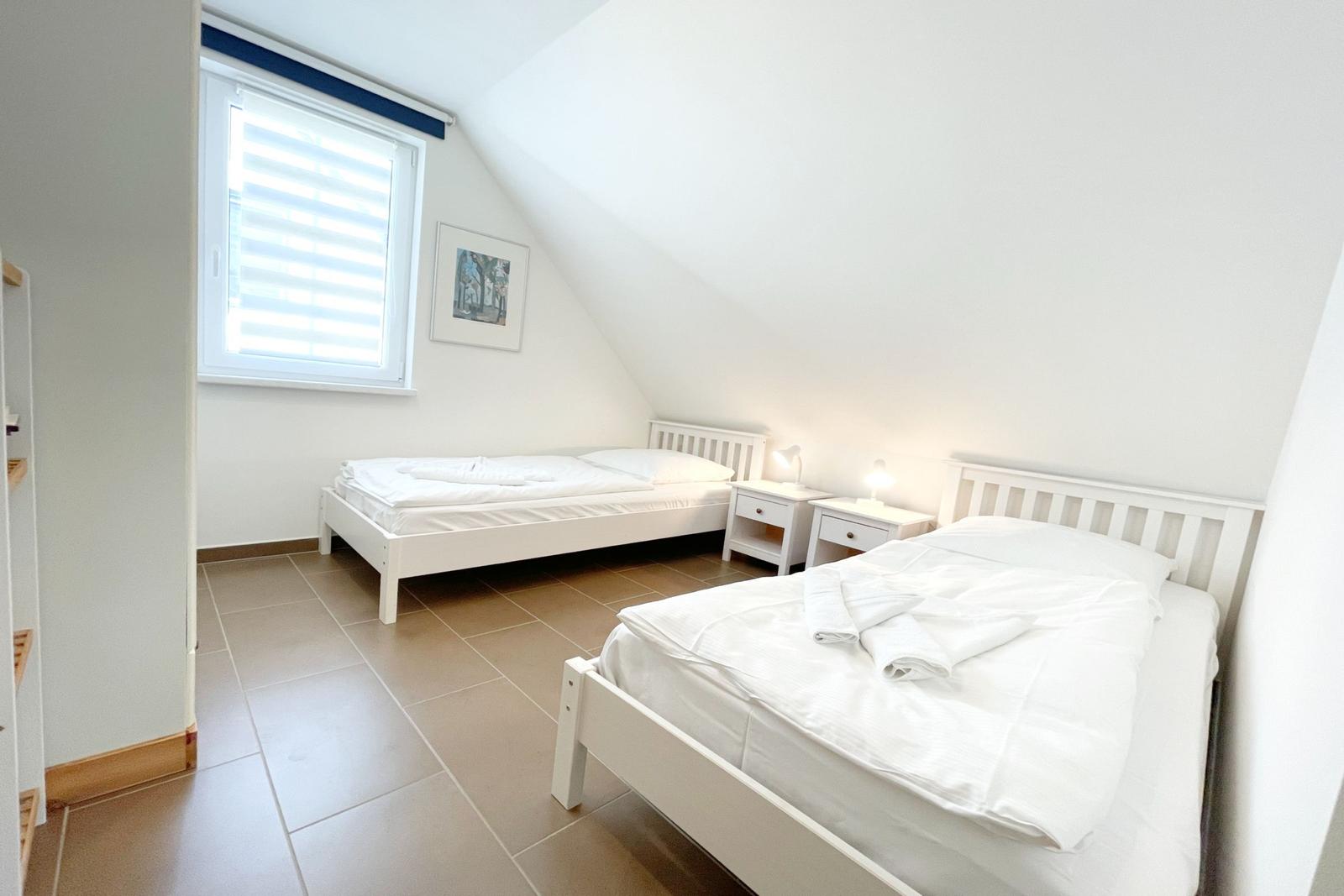 Zwei weiße Betten mit Nachttischen und Fenster im Dachzimmer.