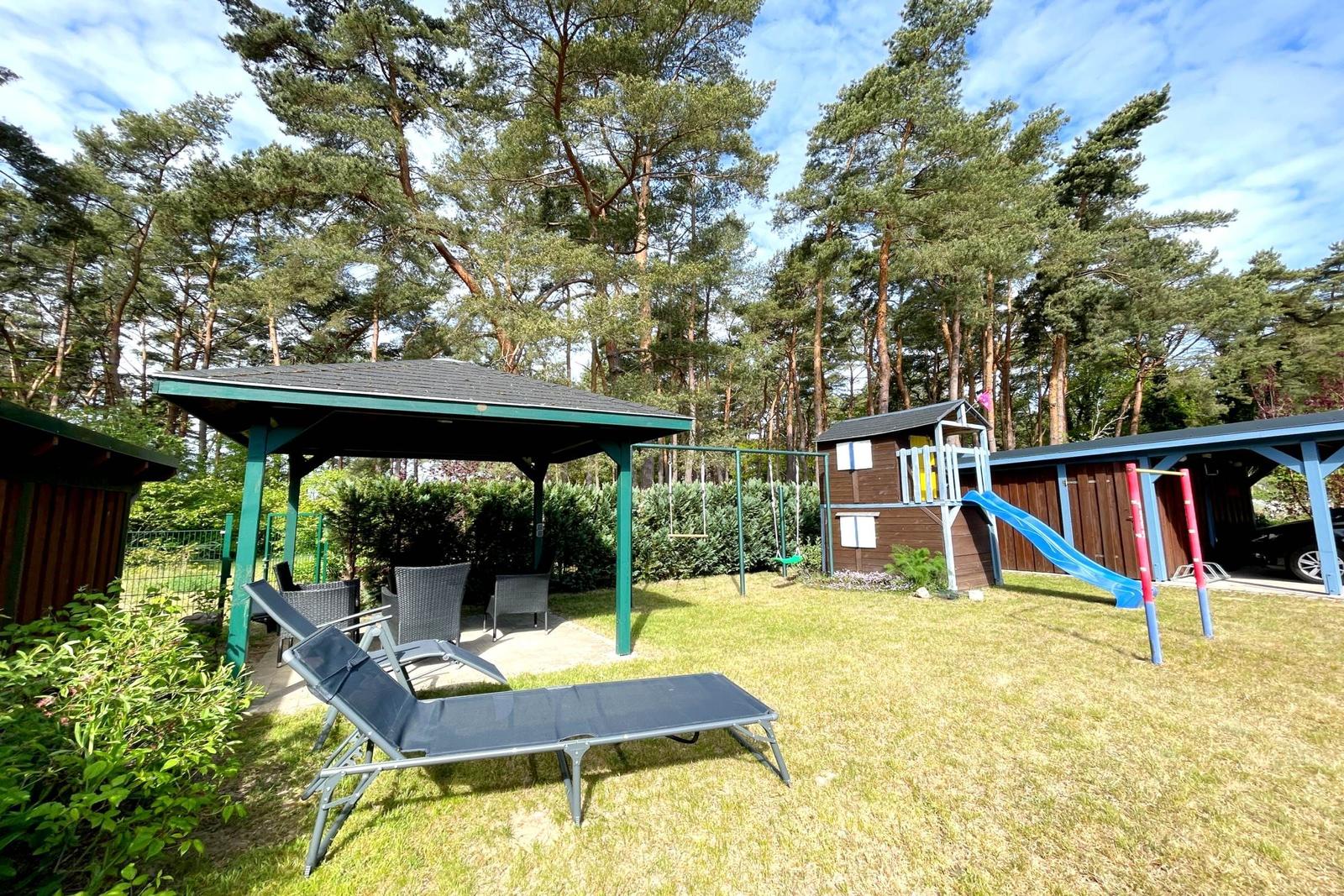 Grüner Garten mit Liegestühlen, Spielplatz und Pavillon unter Bäumen.