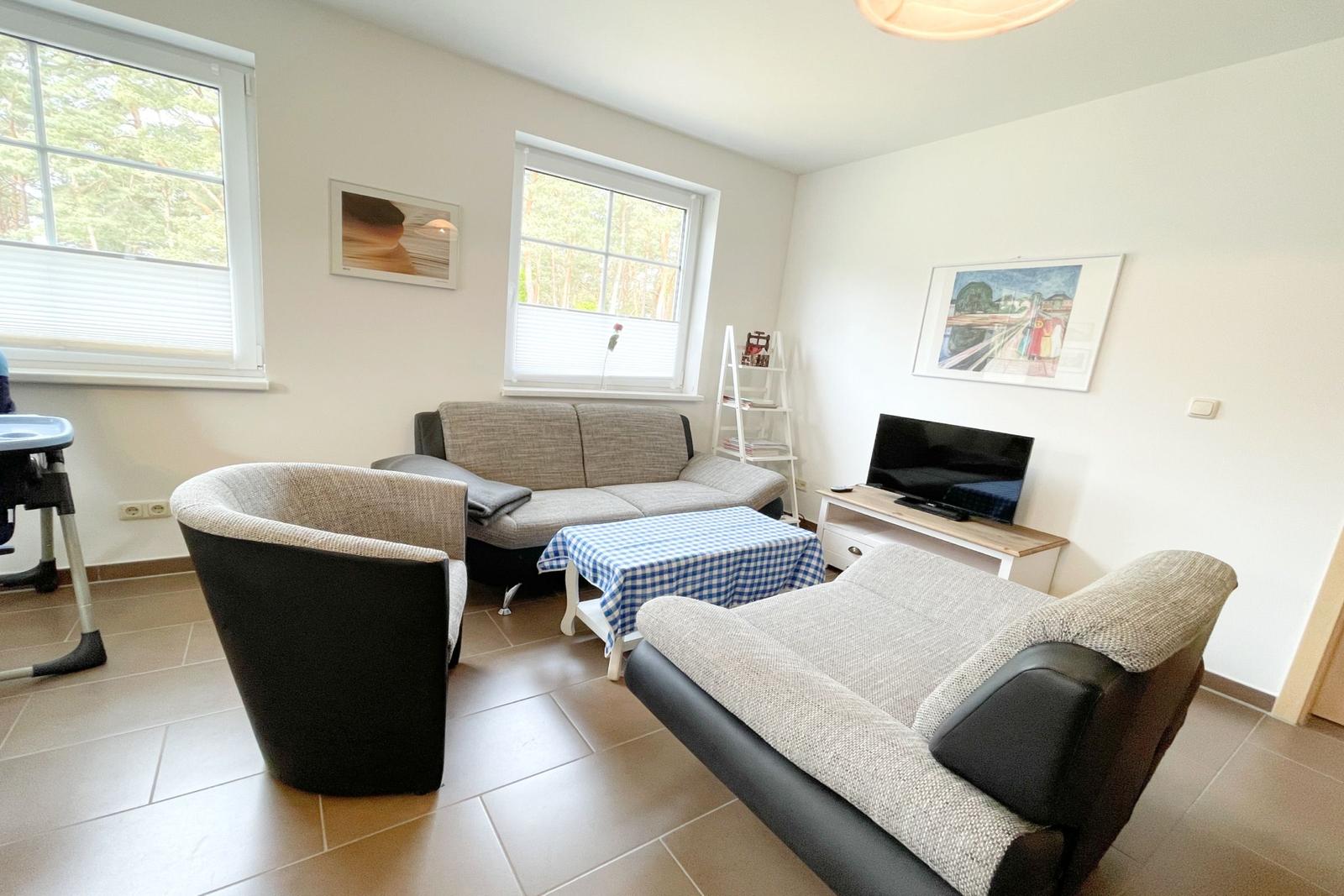 Wohnzimmer mit Sofa, Sessel und Fernseher. Fenster zeigen Wald. Tisch mit kariertem Tischtuch.