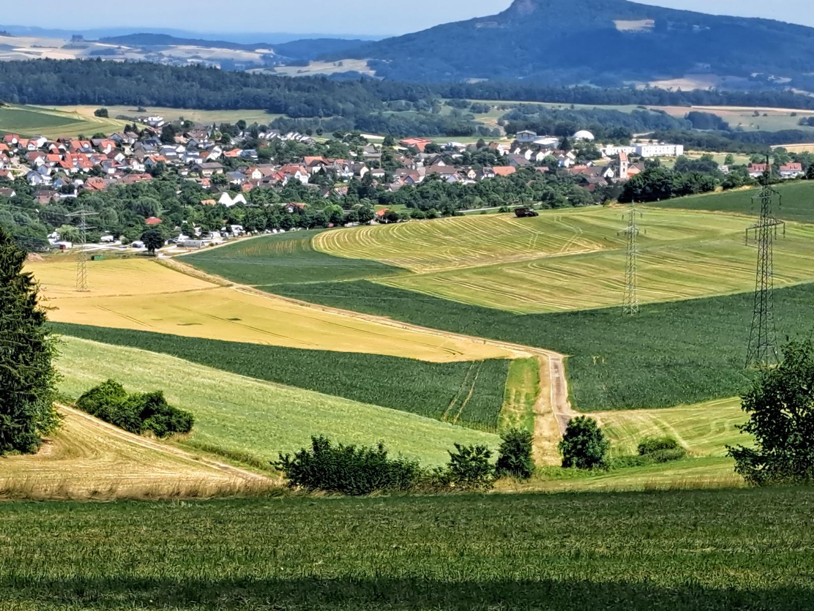 Weitläufige Landschaft mit Feldern, einem Dorf und Bergen im Hintergrund.