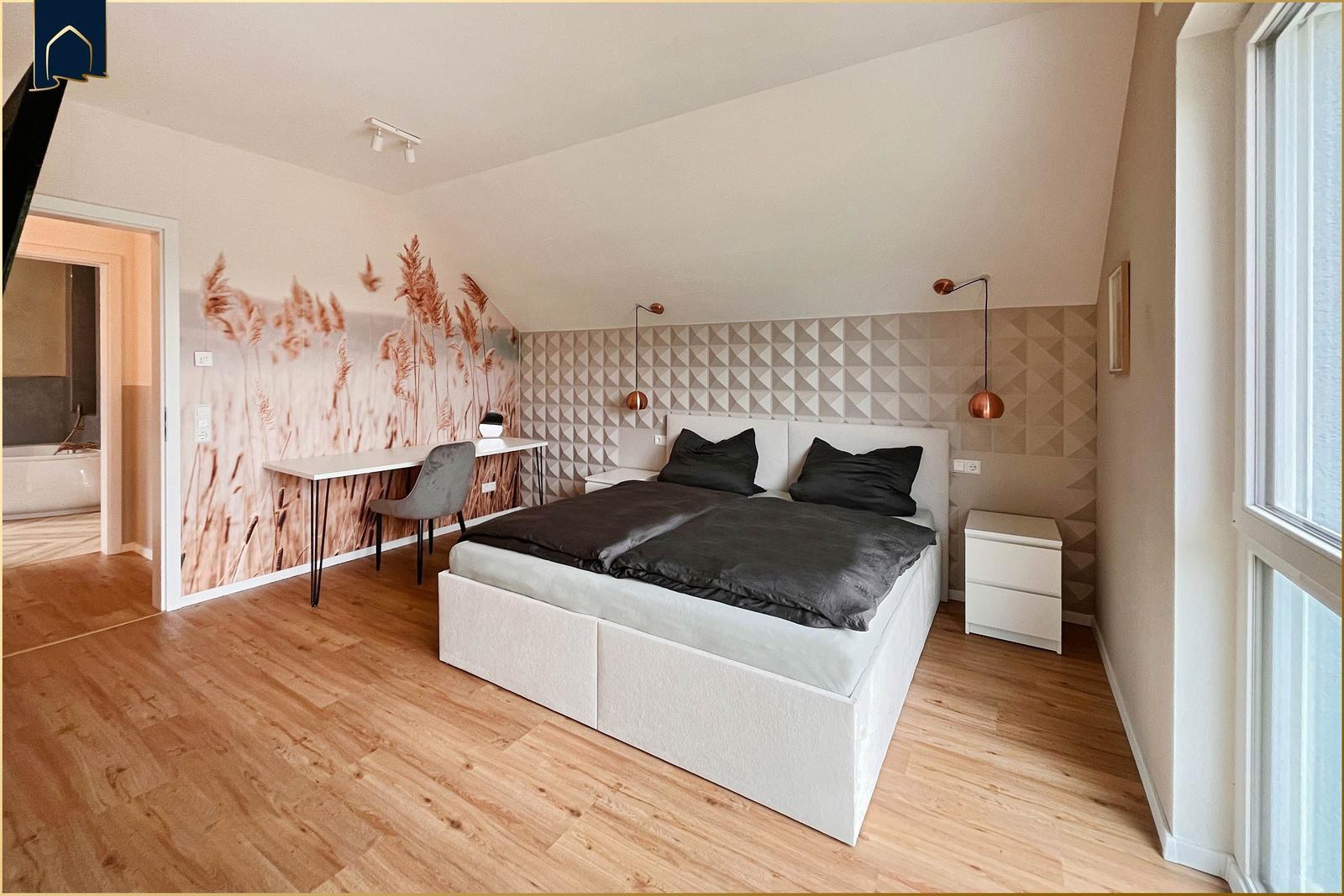 Schlafzimmer mit Doppelbett, Schreibtisch und großem Fenster. Wandtapete mit Pflanzenmotiv.