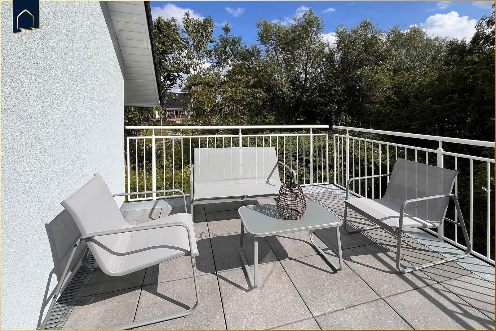 Terrasse mit weißen Sitzmöbeln und Tisch. Blick auf grüne Bäume und einen blauen Himmel.