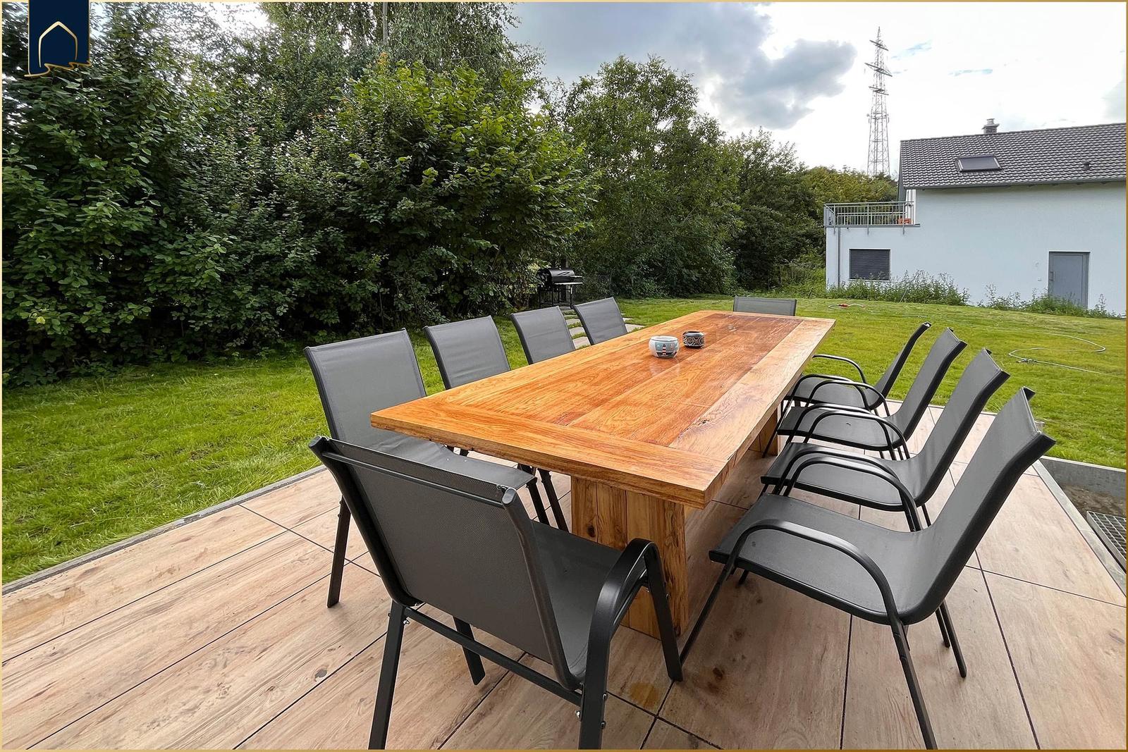 Holztisch mit acht Stühlen auf Terrasse. Grünflächen und Gartenhaus im Hintergrund.