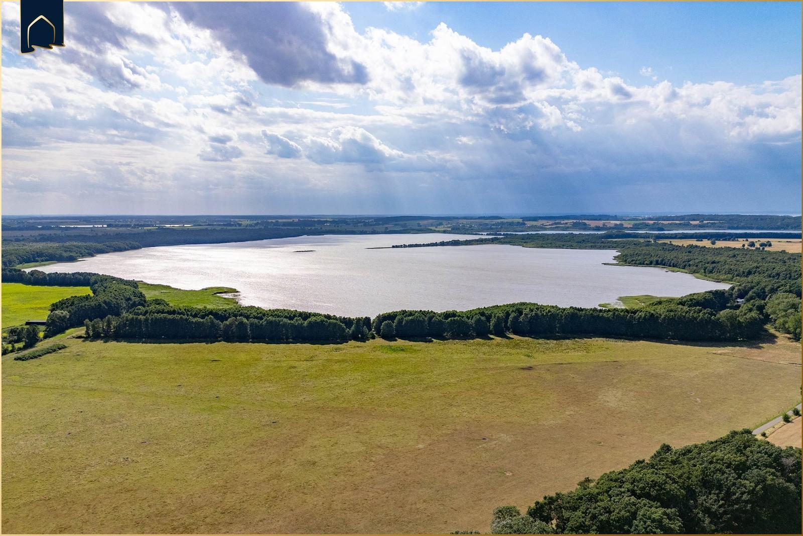 Weitläufiger See mit grünen Wiesen und Waldstreifen unter bewölktem Himmel.