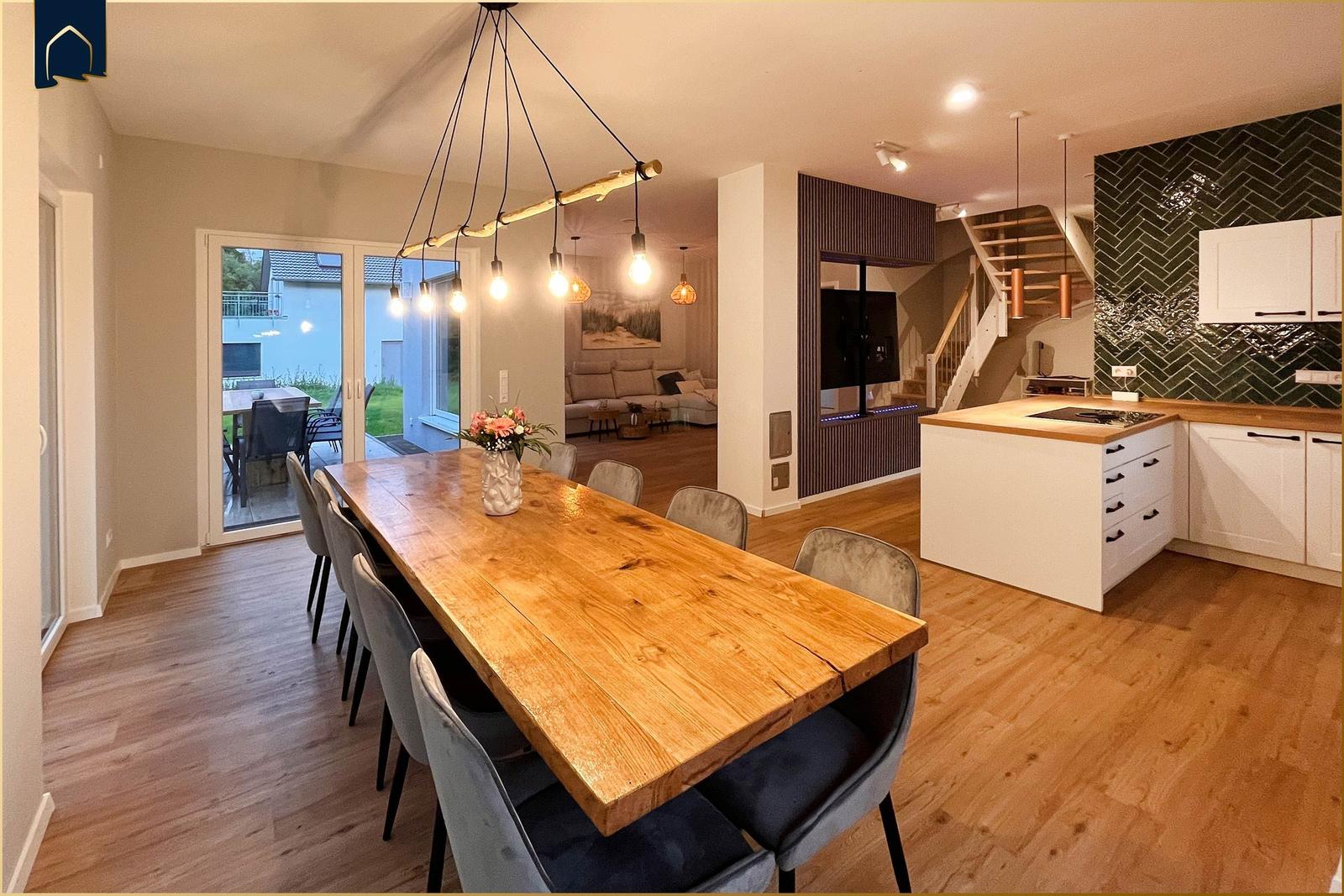 Essbereich mit Holztisch und Stühlen. Küche mit Arbeitsplatte und Wandfliesen. Wohnzimmer im Hintergrund.