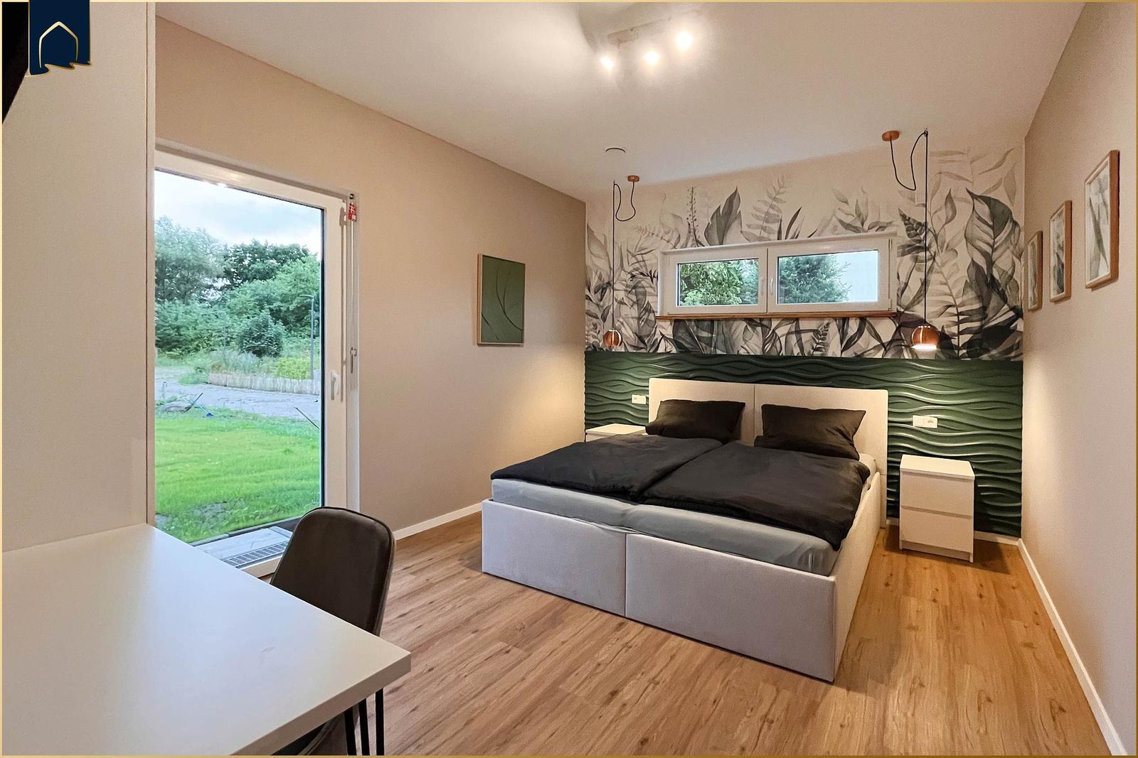Schlafzimmer mit Bett, Schreibtisch und Fenster zur grünen Außenseite.