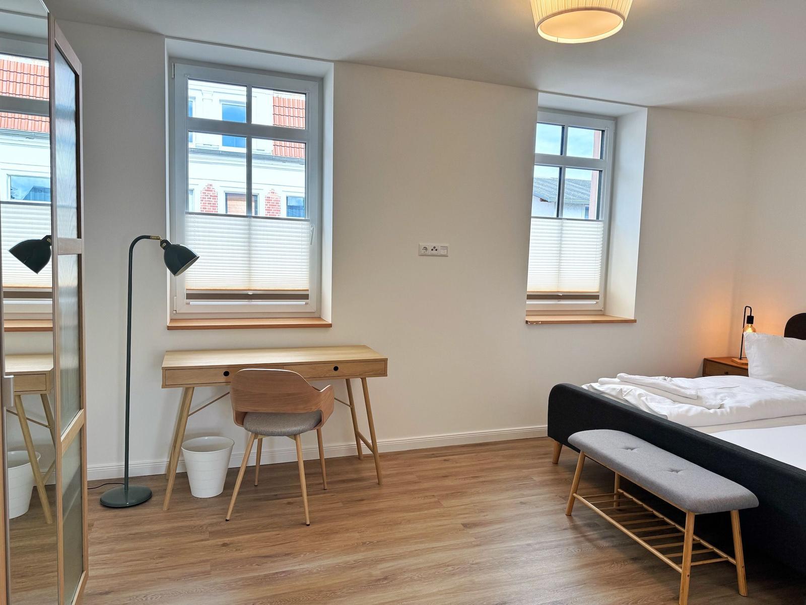 Schlafzimmer mit Bett, Schreibtisch und zwei Fenstern.