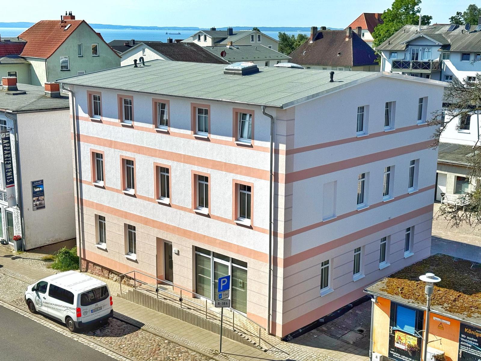 Zweistöckiges Gebäude mit rosa Streifen und Fenstern. Parkplatz und Straße sind sichtbar.