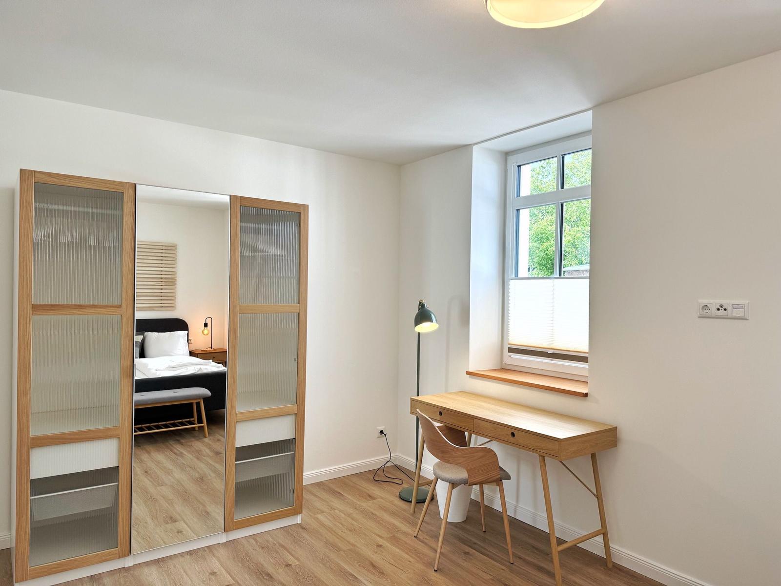 Schlafzimmer mit Schrank und Schreibtisch. Fenster mit Jalousie und Stehlampe.