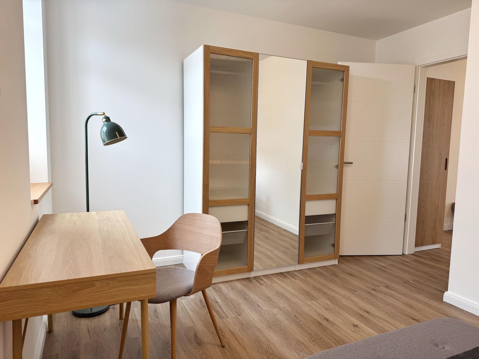 Schreibtisch mit Stuhl und Stehlampe neben Kleiderschrank mit Spiegeltüren.