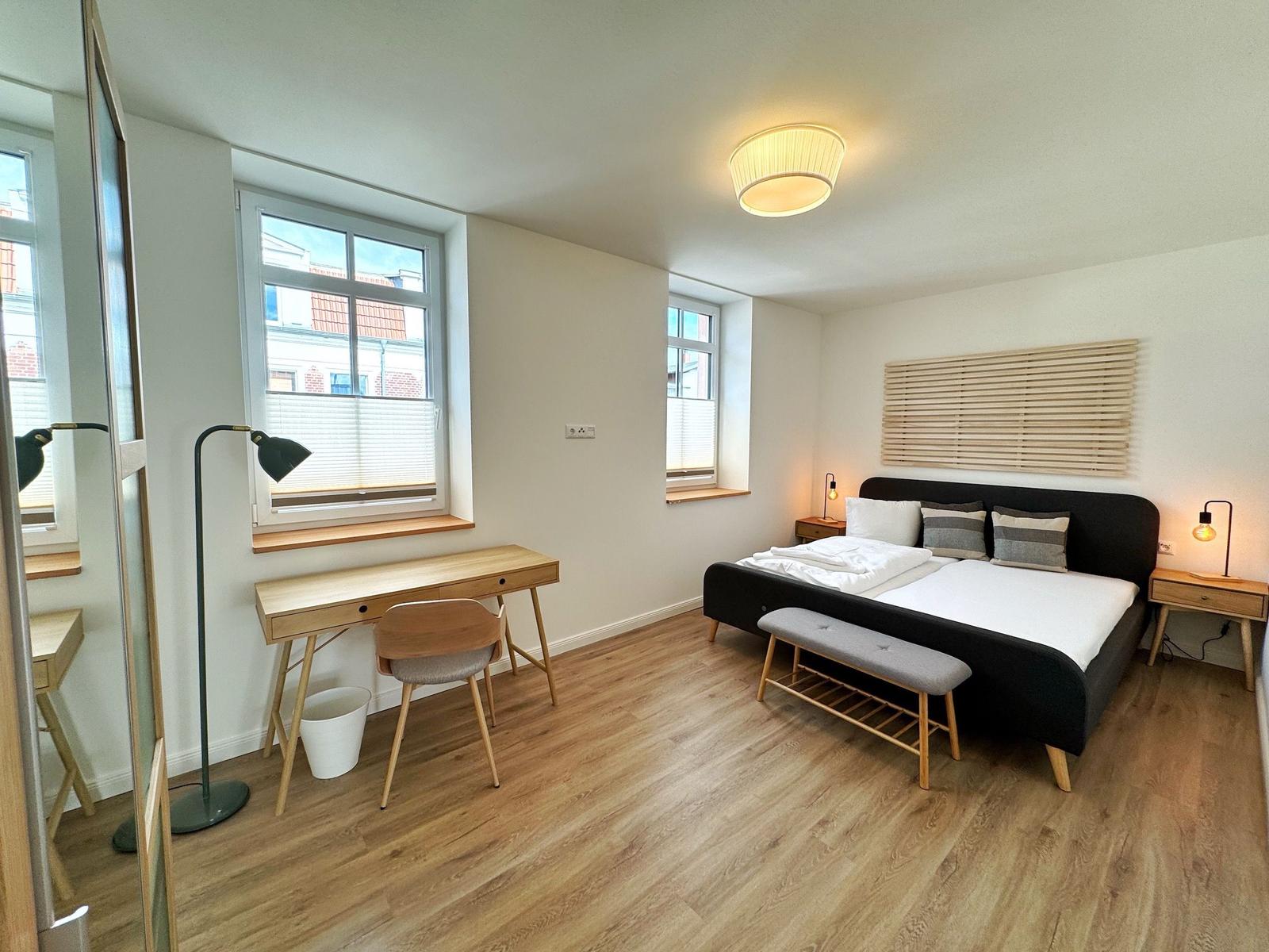 Schlafzimmer mit Bett, Schreibtisch und zwei Fenstern.