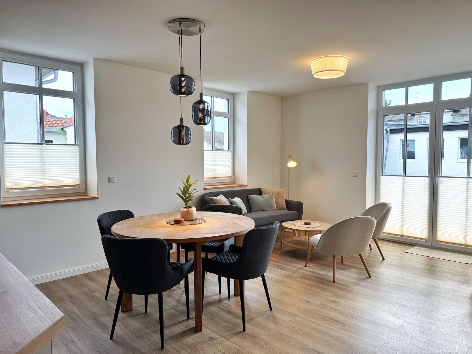 Essbereich mit runder Tisch und Stühlen, Wohnbereich mit Sofa und Sesseln, große Fenster und Holzboden.