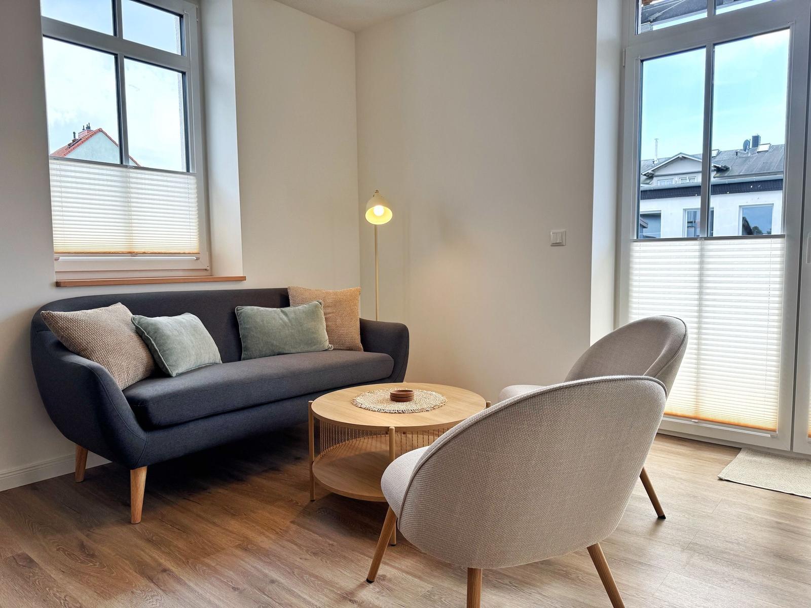 Ein Wohnzimmer mit Sofa, zwei Stühlen und einem runden Tisch. Große Fenster mit Jalousien.