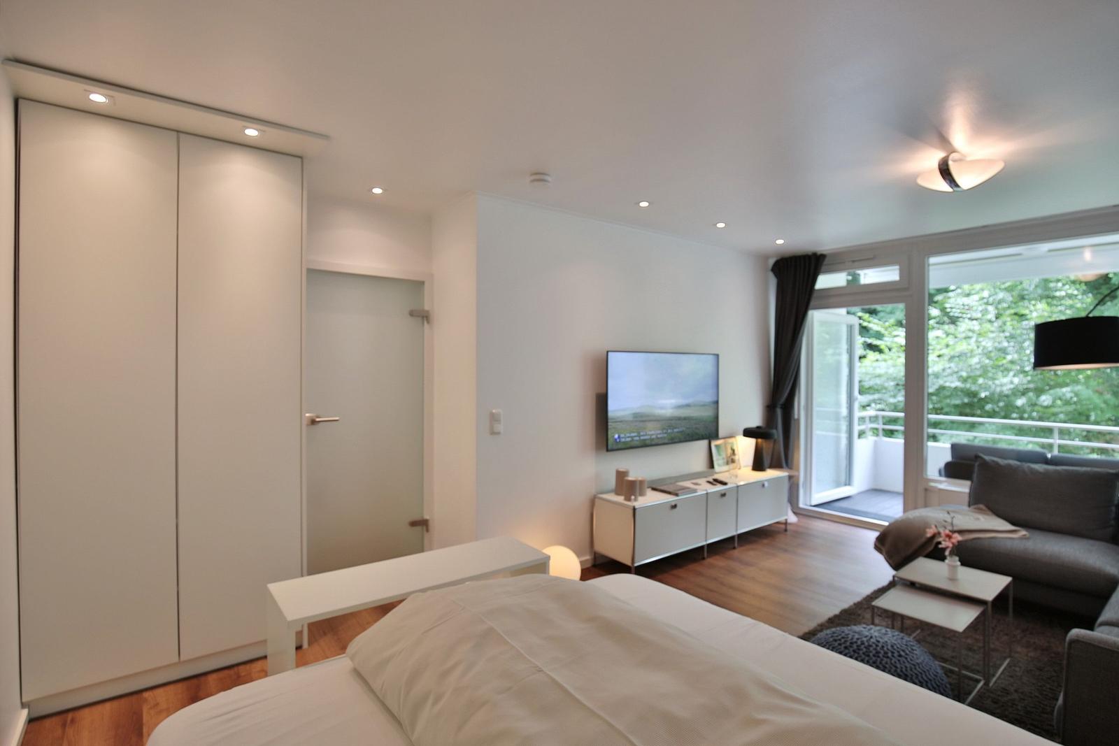 Schlafzimmer mit Bett, Kleiderschrank und TV. Wohnbereich mit Sofa und Balkon.