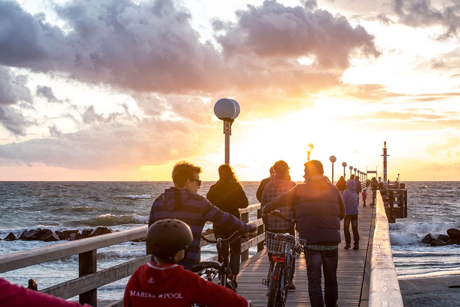Besucher genießen Sonnenuntergang auf Holzsteg über Meer mit Fahrrädern.
