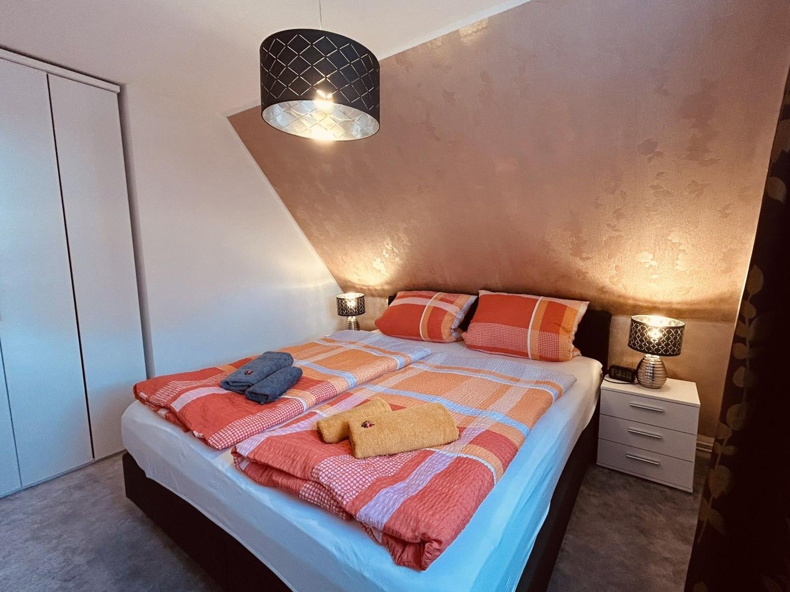 Schlafzimmer mit Bett, Nachttisch und Kleiderschrank. Decke und Kissen sind rot-orange kariert.