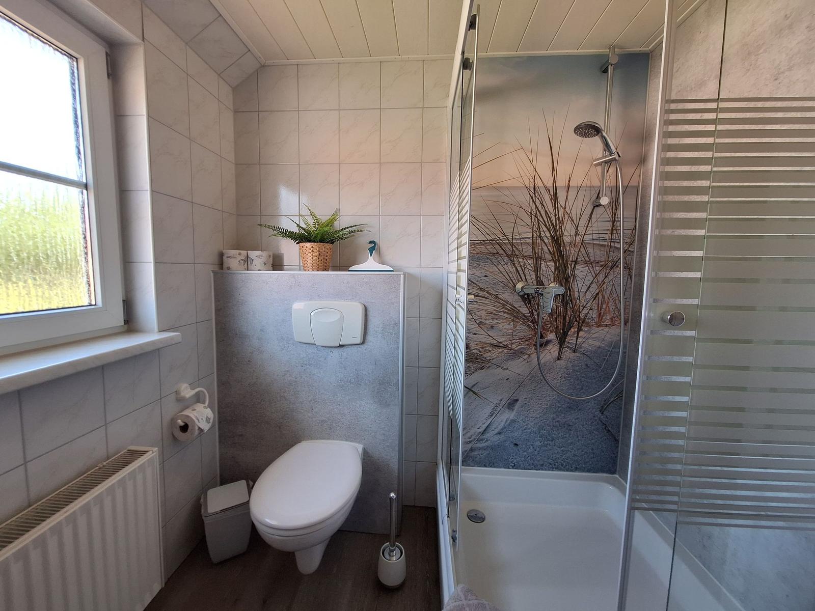 Bad mit Dusche, WC und Fenster. Duschwand zeigt Strandmotiv.