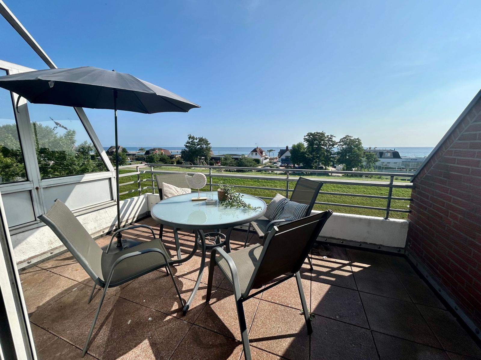 Terrasse mit Tisch, Stühlen und Sonnenschirm. Blick auf Grünfläche und Meer.