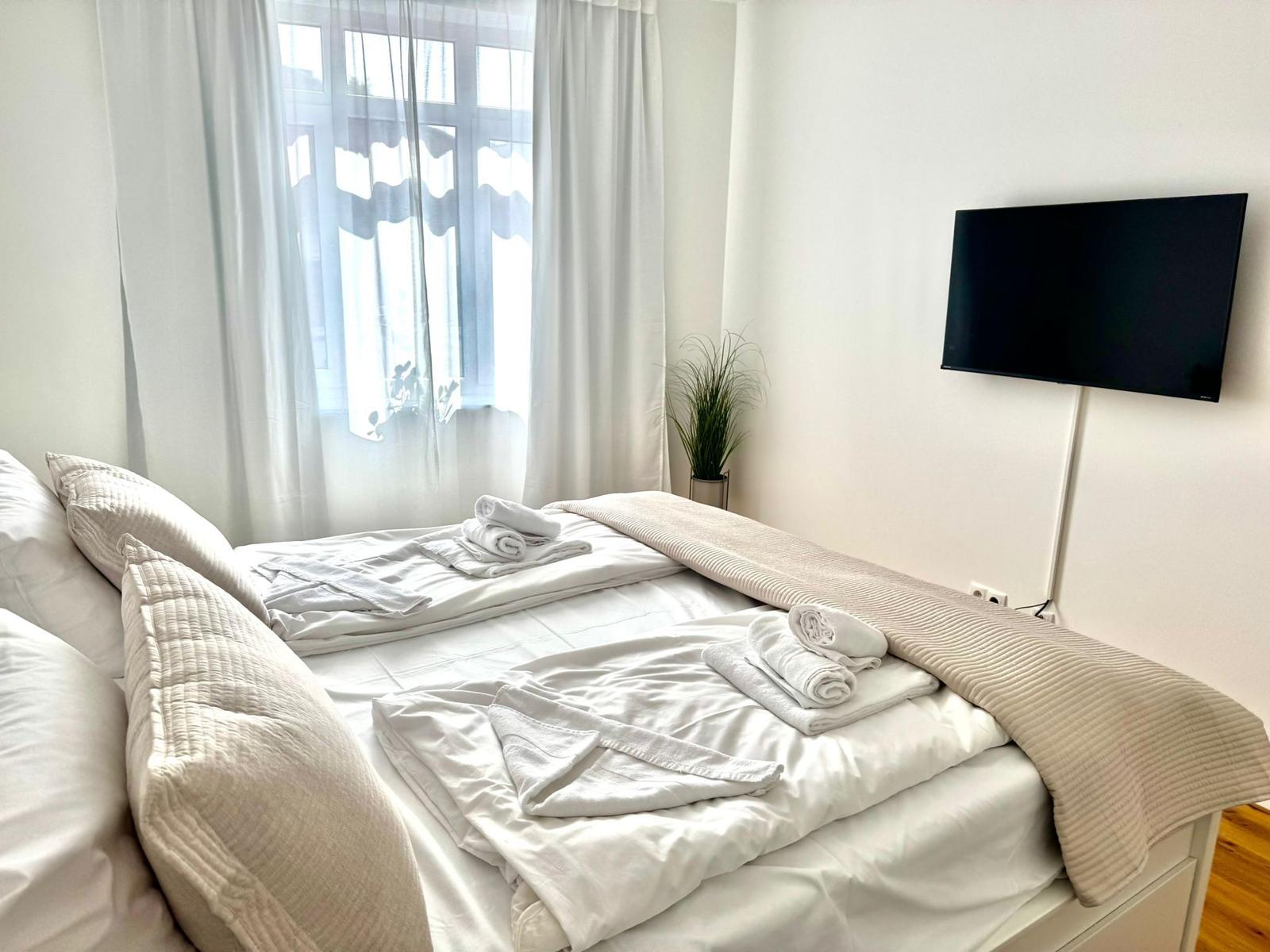 Schlafzimmer mit Bett, TV und Fenster mit weißen Vorhängen.