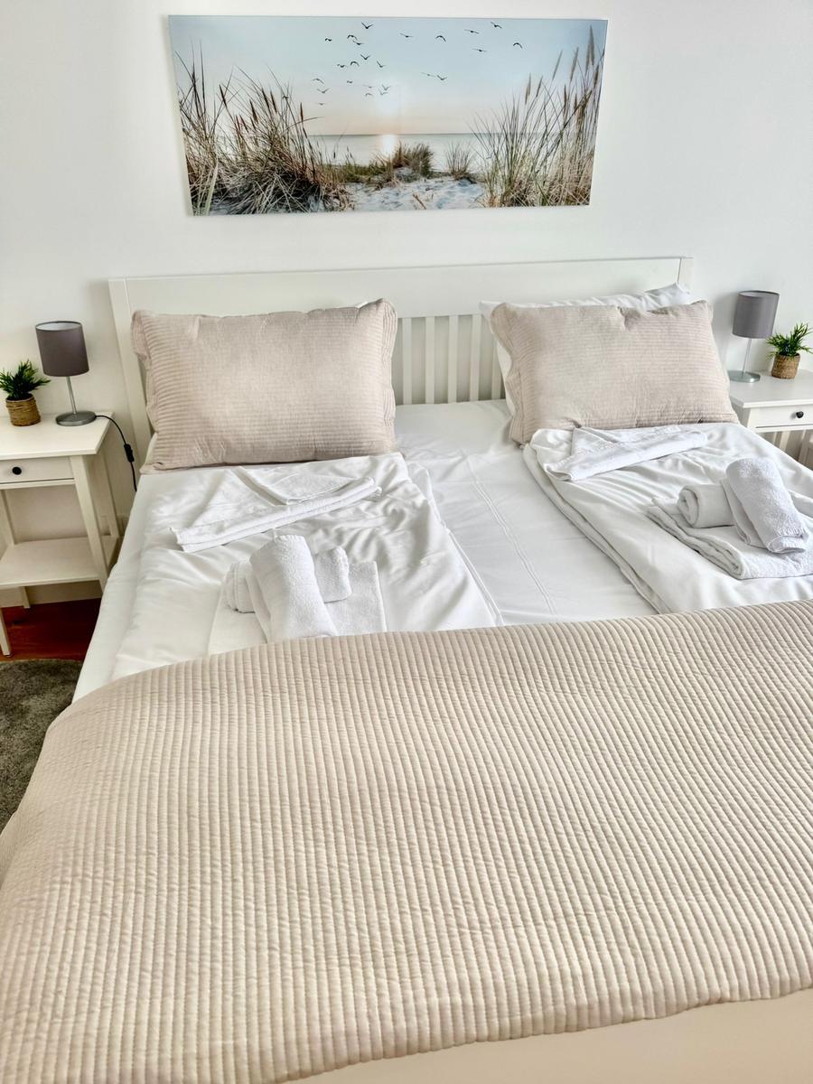 Doppelbett mit weißer Bettwäsche und hellen Kissen. An den Seiten Nachttische mit Lampen.