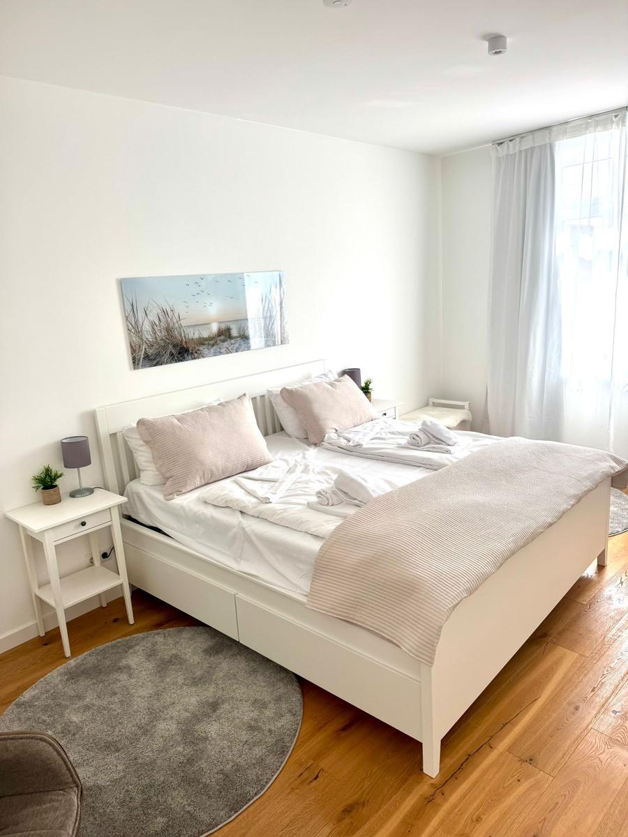 Ein Schlafzimmer mit weißem Bett, Holzboden und Fenster mit weißen Vorhängen.