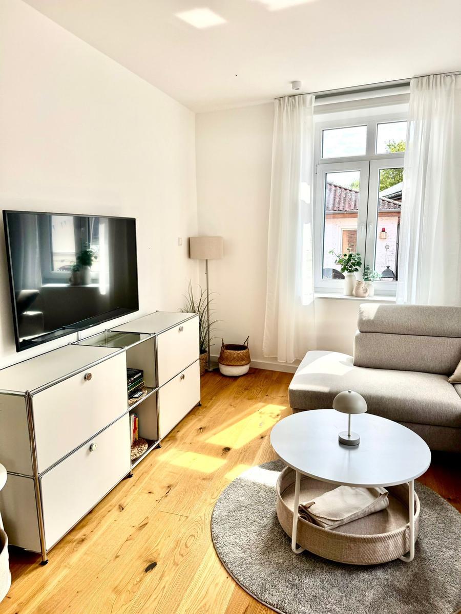 Wohnzimmer mit Sofa, TV und Fenster. Holzboden und runder Tisch.