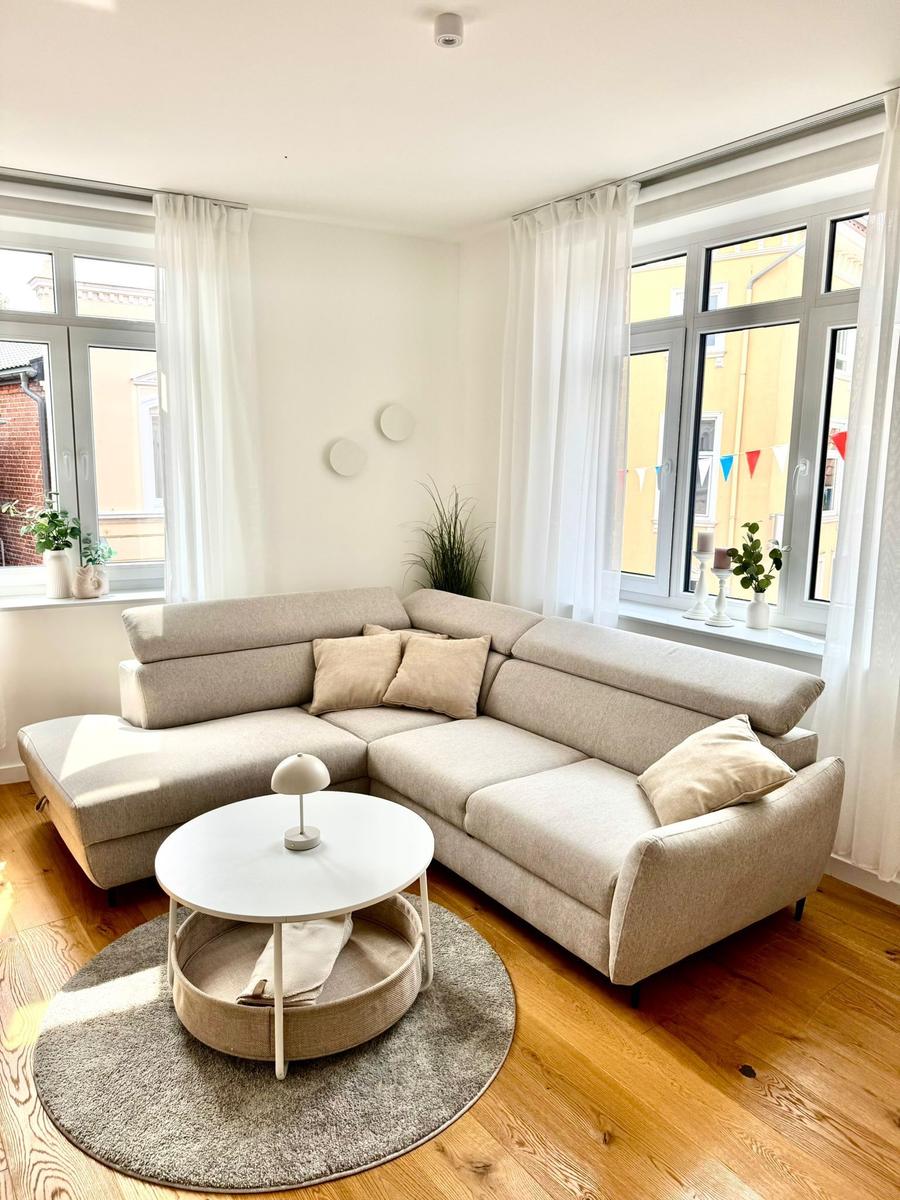 Wohnzimmer mit Ecksofa, runden Tisch und Holzboden. Große Fenster mit weißen Vorhängen.