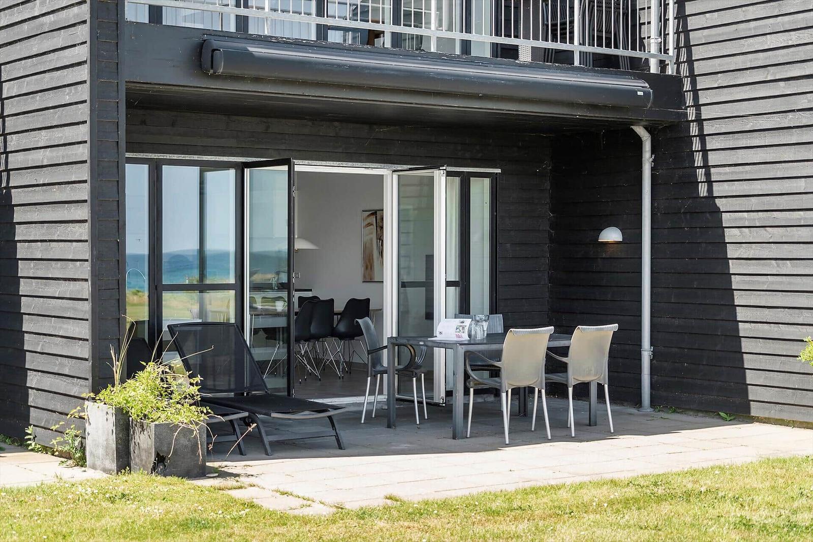 Terrasse mit Tisch, Stühlen und Liegestuhl. Durchgehende Glastüren führen ins Haus.