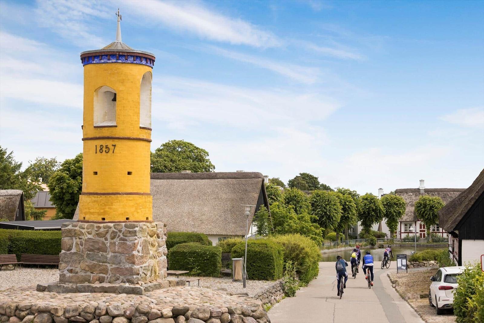 Gelber Turm mit Jahreszahl 1857, Radfahrer auf Weg zwischen Häusern mit Strohdächern.