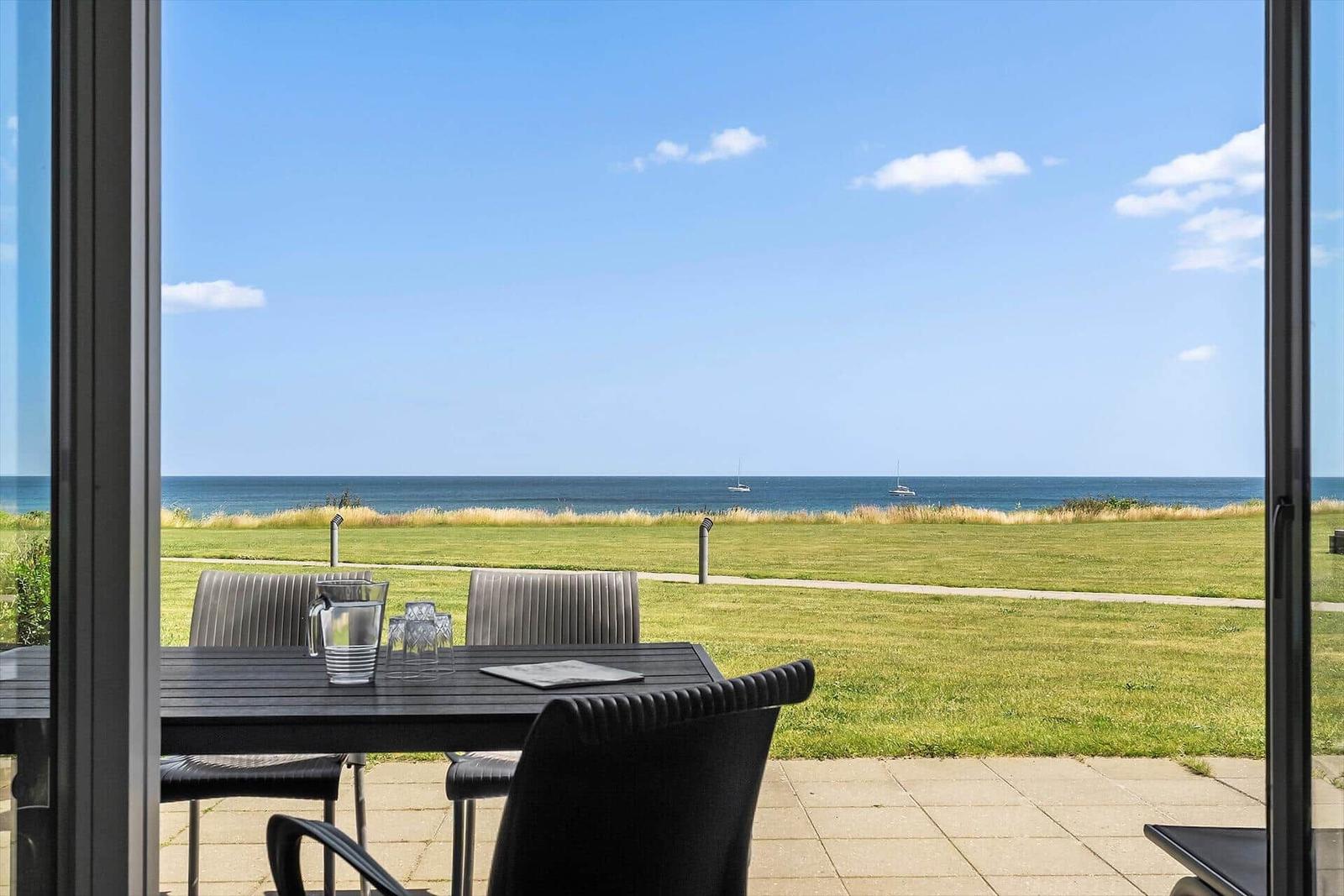 Terrasse mit Tisch und Stühlen, Blick auf Grasfläche und Meer mit Booten.