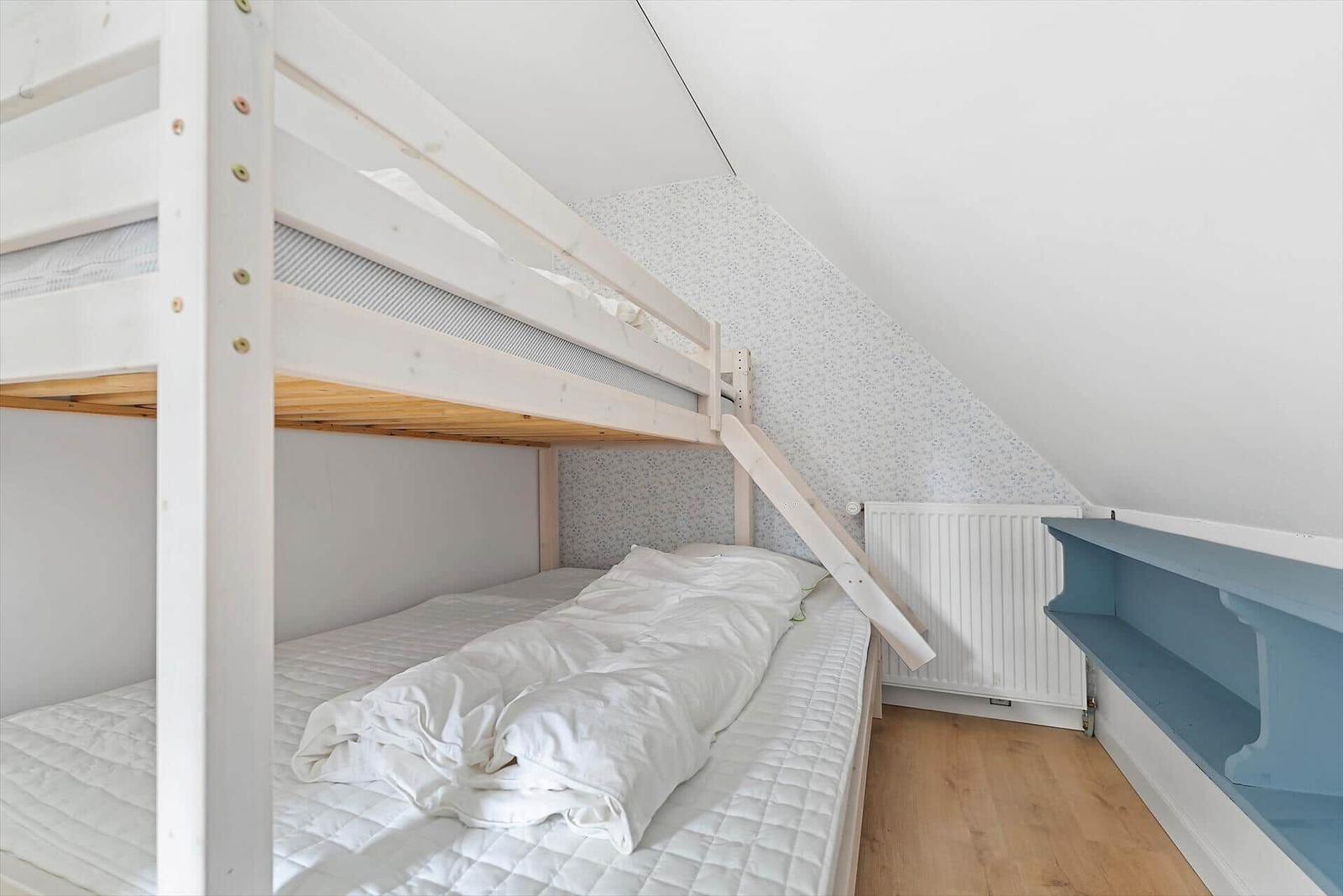 Doppelstockbett mit Matratzen und weißer Bettwäsche in einem Dachzimmer mit Holzboden und Regal.