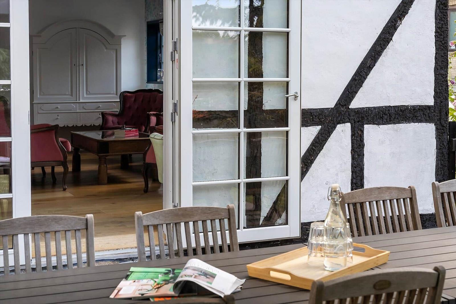 Terrasse mit Tisch und Stühlen. Durch Glastüren ist Wohnzimmer mit Möbeln sichtbar.