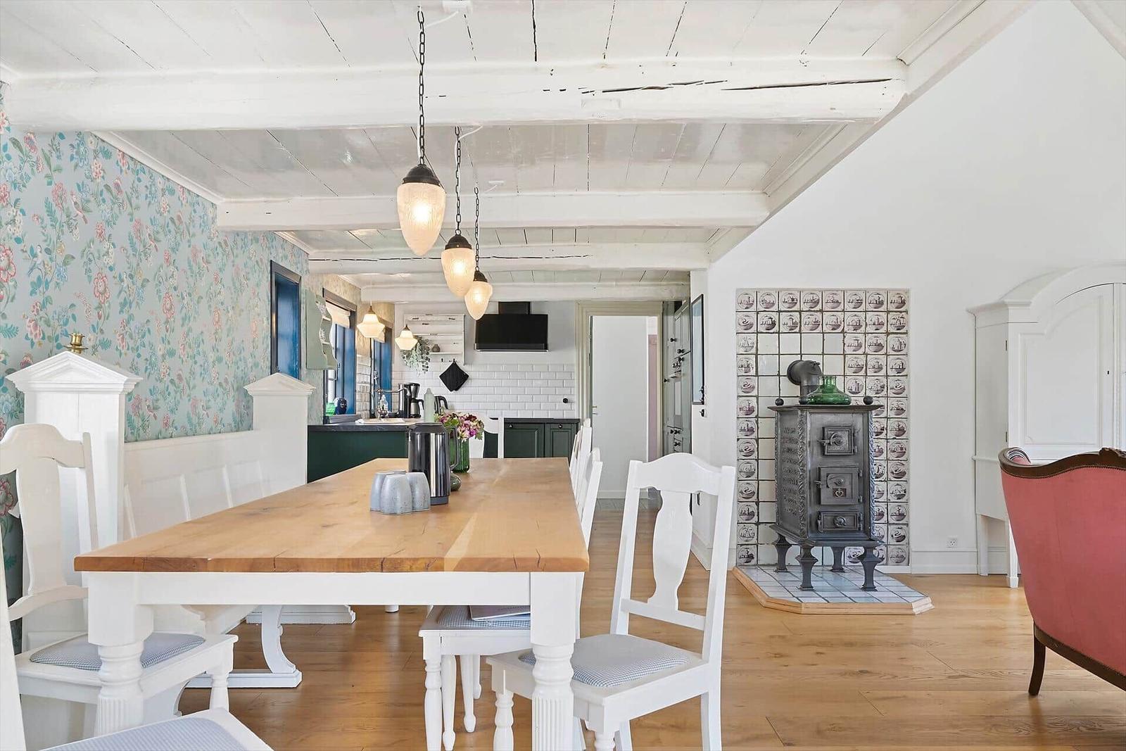 Esszimmer mit Holztisch, weißen Stühlen und Kachelofen an der Wand.