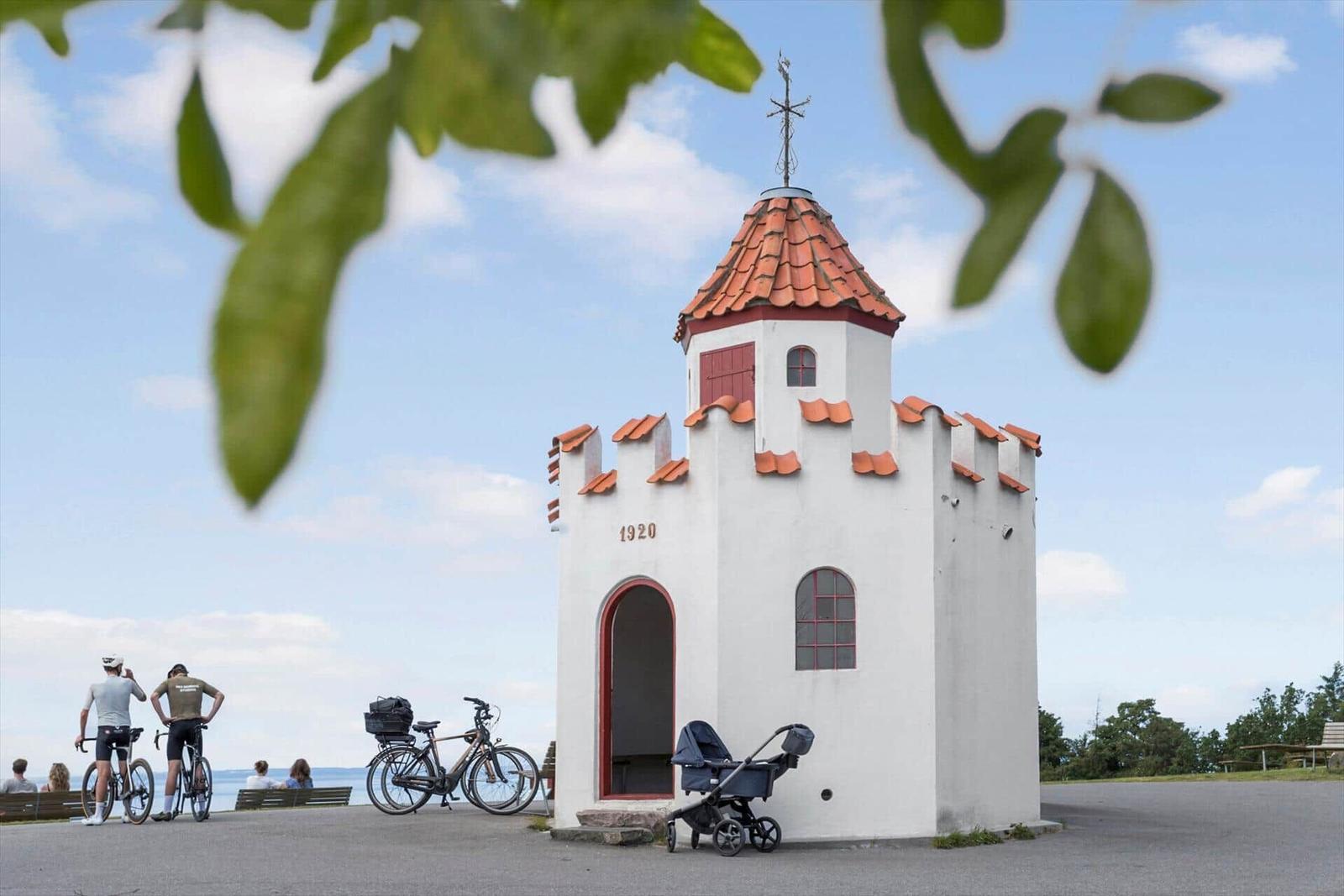 Kleine weiße Kapelle mit rotem Dach und Tür. Fahrräder und Kinderwagen stehen daneben.