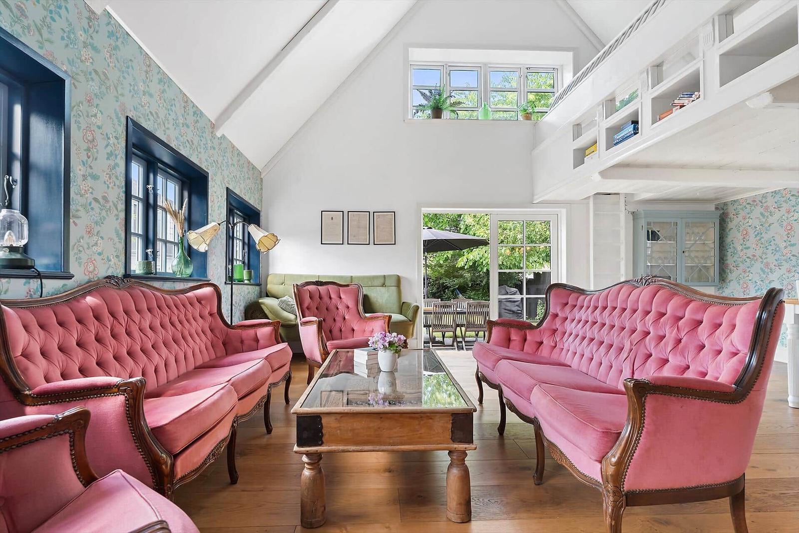 Wohnzimmer mit rosa Polstersofas, Glas-Tisch und Holzboden. Fenster mit floraler Tapete.