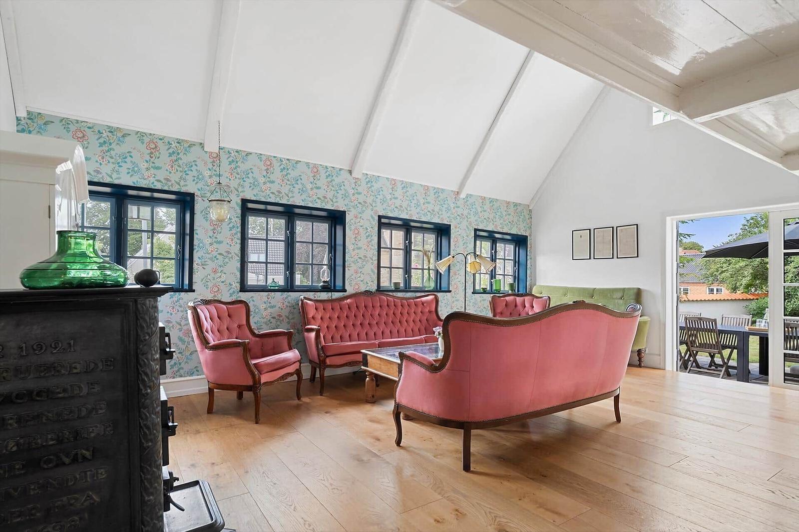 Wohnzimmer mit rosa Sitzmöbeln, floraler Tapete und Holzboden. Durchgang zur Terrasse.