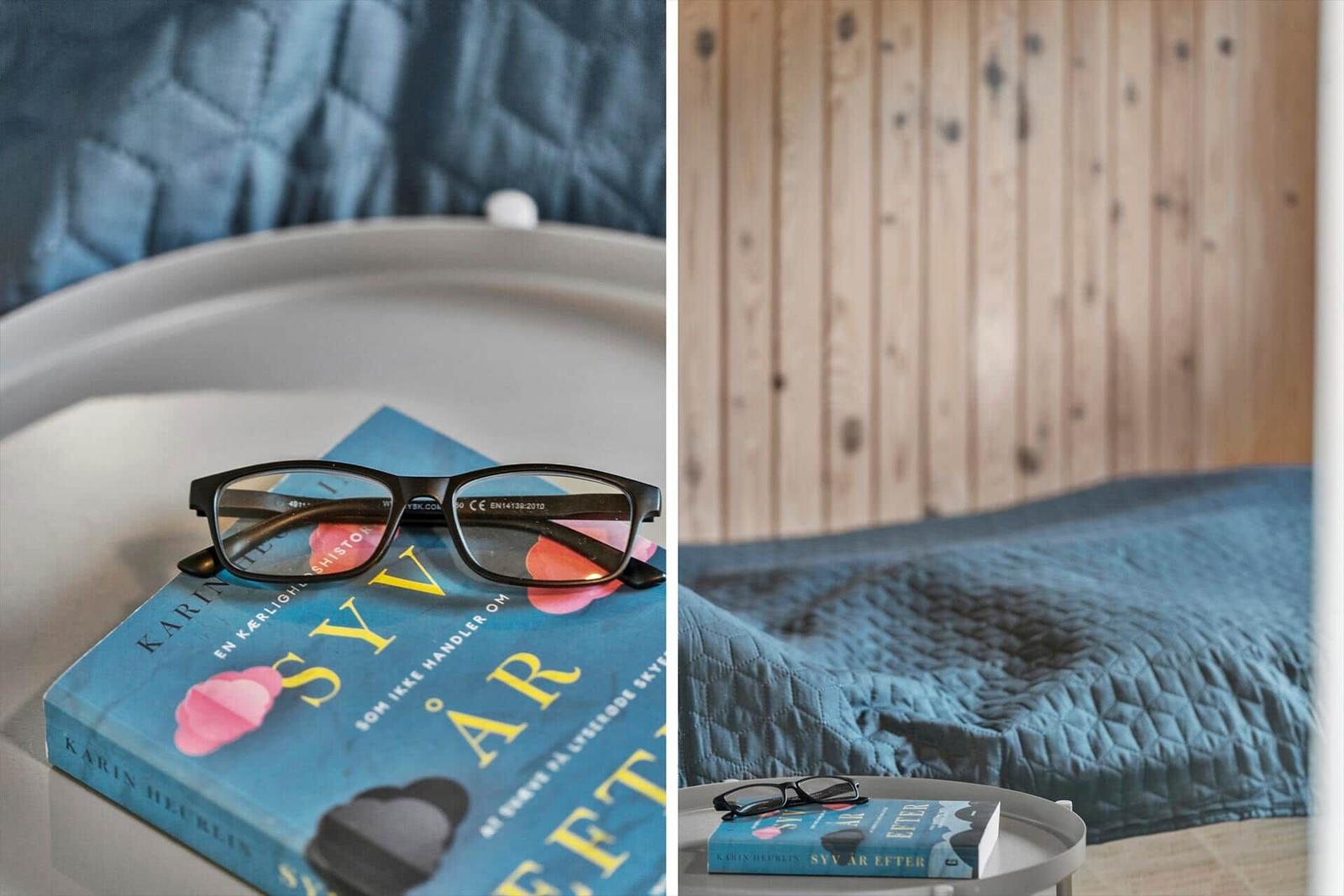 Ein Buch und eine Brille auf einem Tisch neben einem Bett mit blauer Bettdecke.