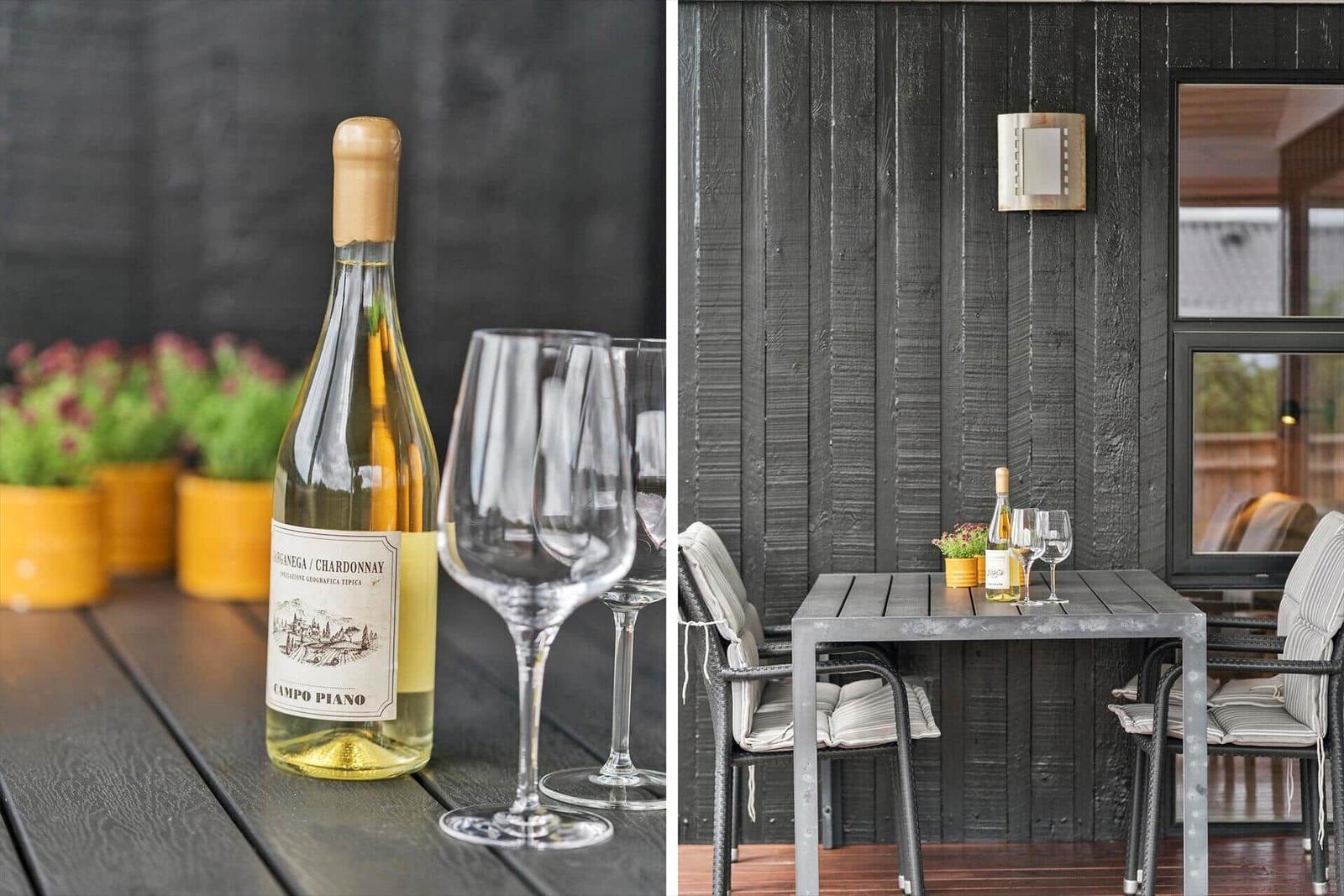 Ein Tisch mit Weinflasche und Gläsern auf einer Terrasse mit dunkler Holzwand.