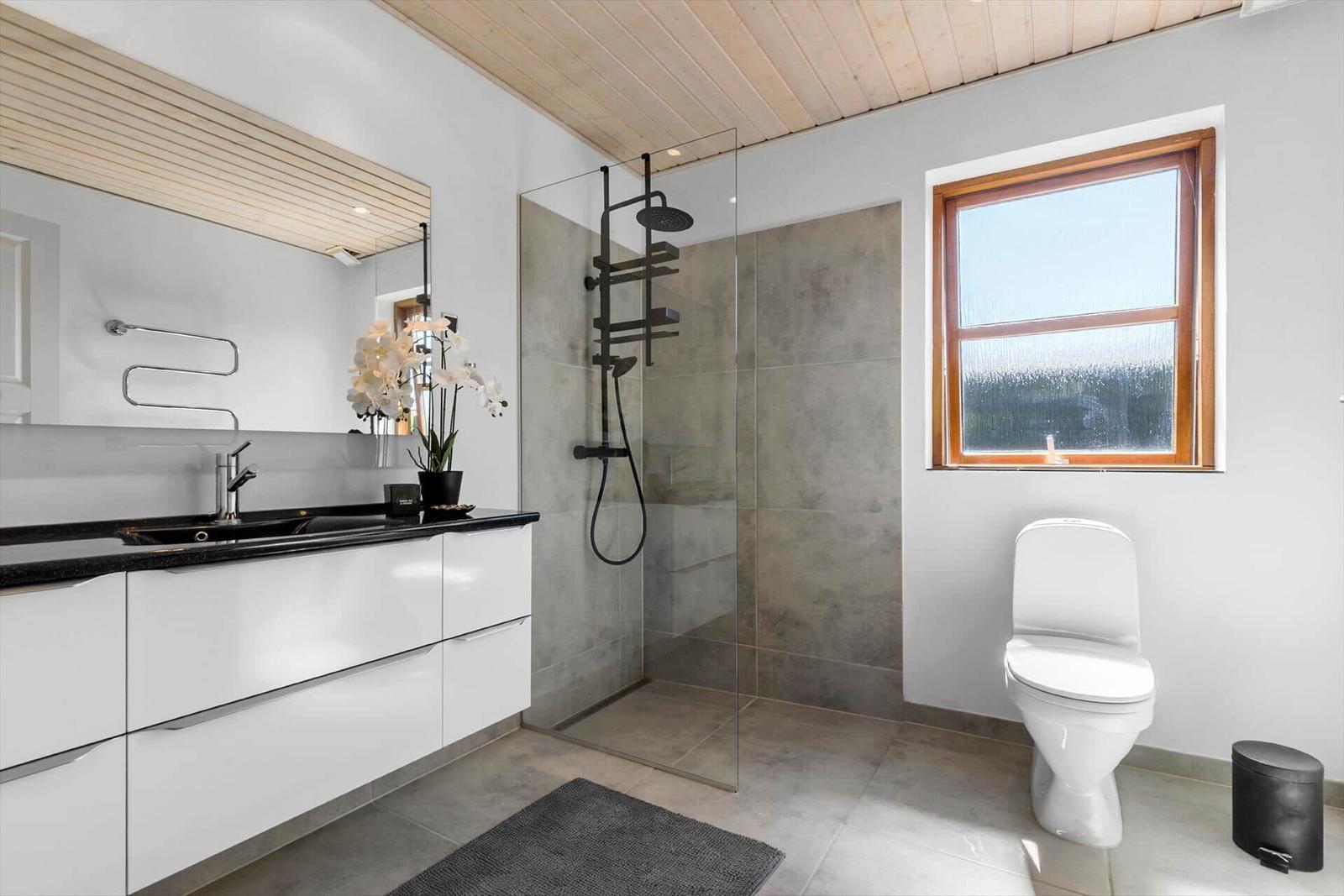 Badezimmer mit Dusche, Waschbecken und Toilette. Holzdecke und Fenster mit Holzrahmen.