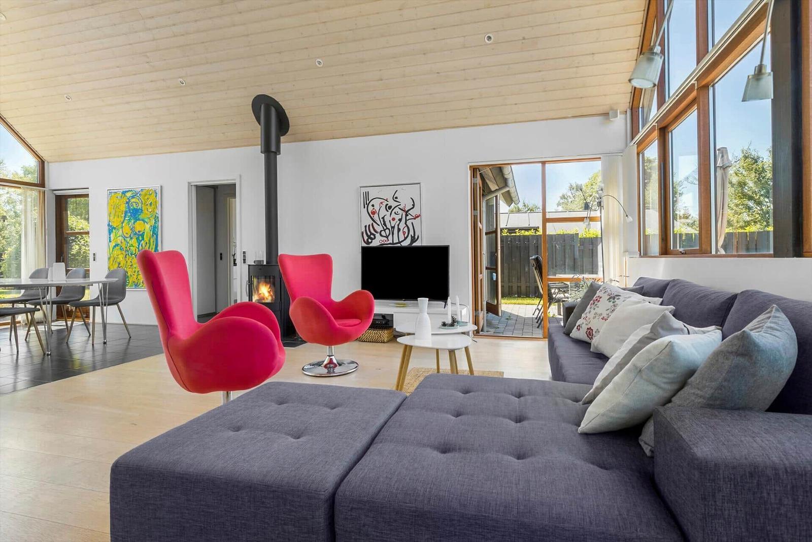 Wohnzimmer mit grauem Sofa, roten Stühlen und Kamin. Große Fenster und Türen zu Garten.